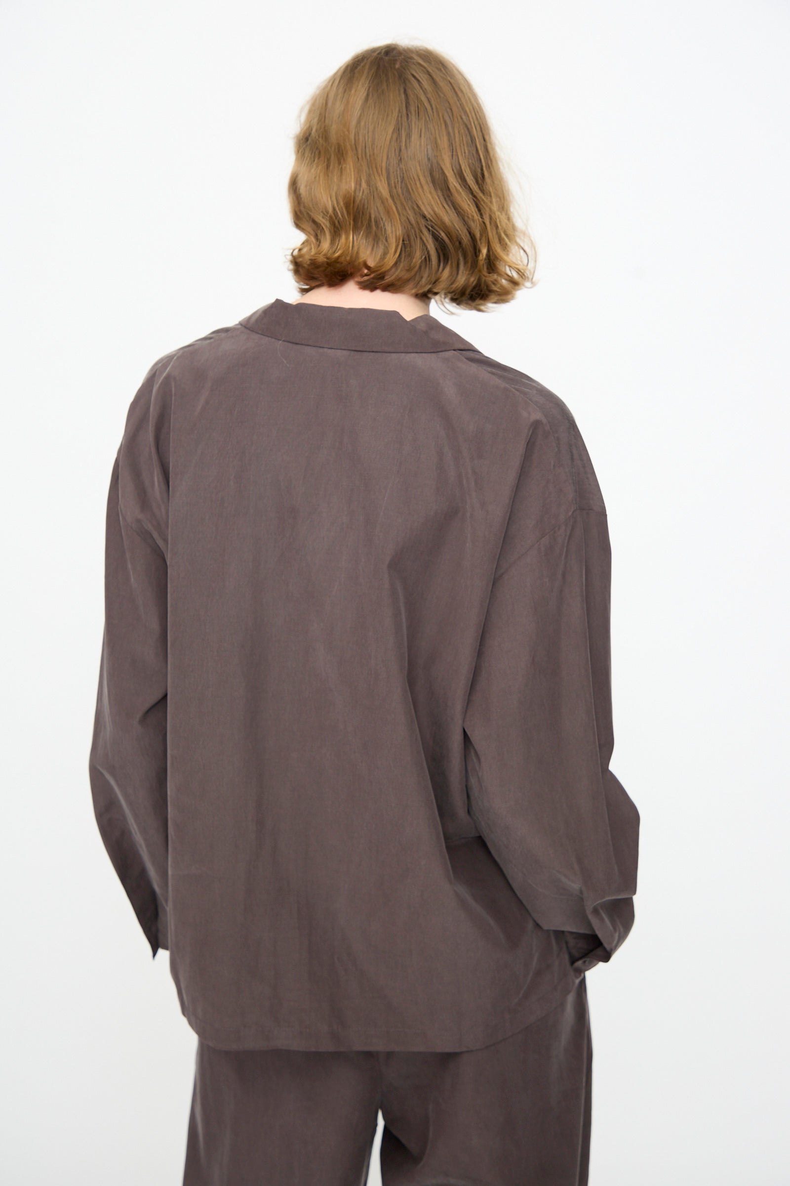 A person with shoulder-length light brown hair is seen from the back against a plain white background, wearing Black Crane’s Cropped Shirt in Dark Plum—a loose-fitting, boxy long-sleeved top crafted from a soft cotton cupro blend.