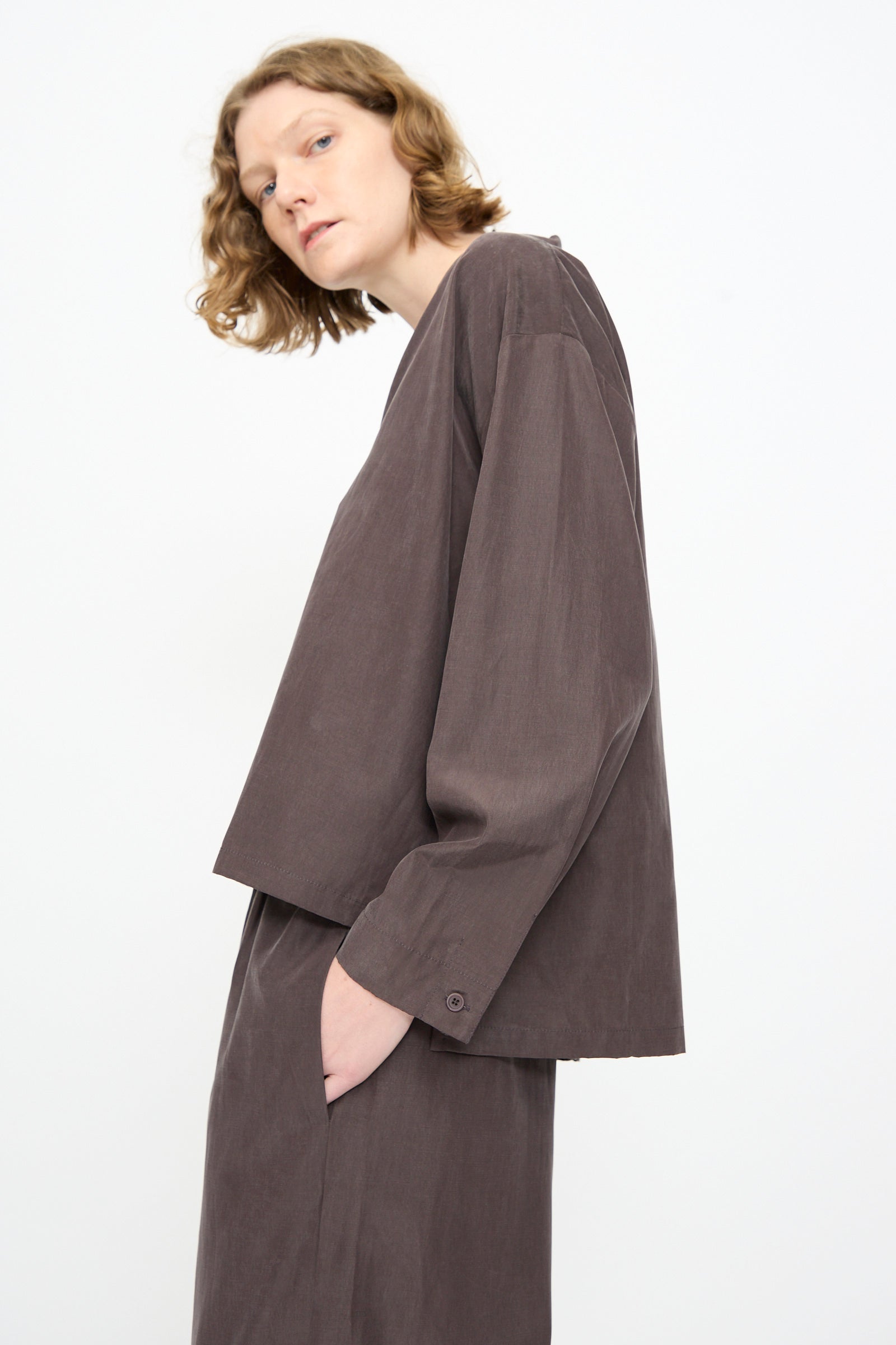 A person with short wavy hair wears the Black Crane Cropped Shirt in Dark Plum—a boxy-fit, long-sleeved top crafted from a soft cotton cupro blend—with matching pants, standing against a plain white background.
