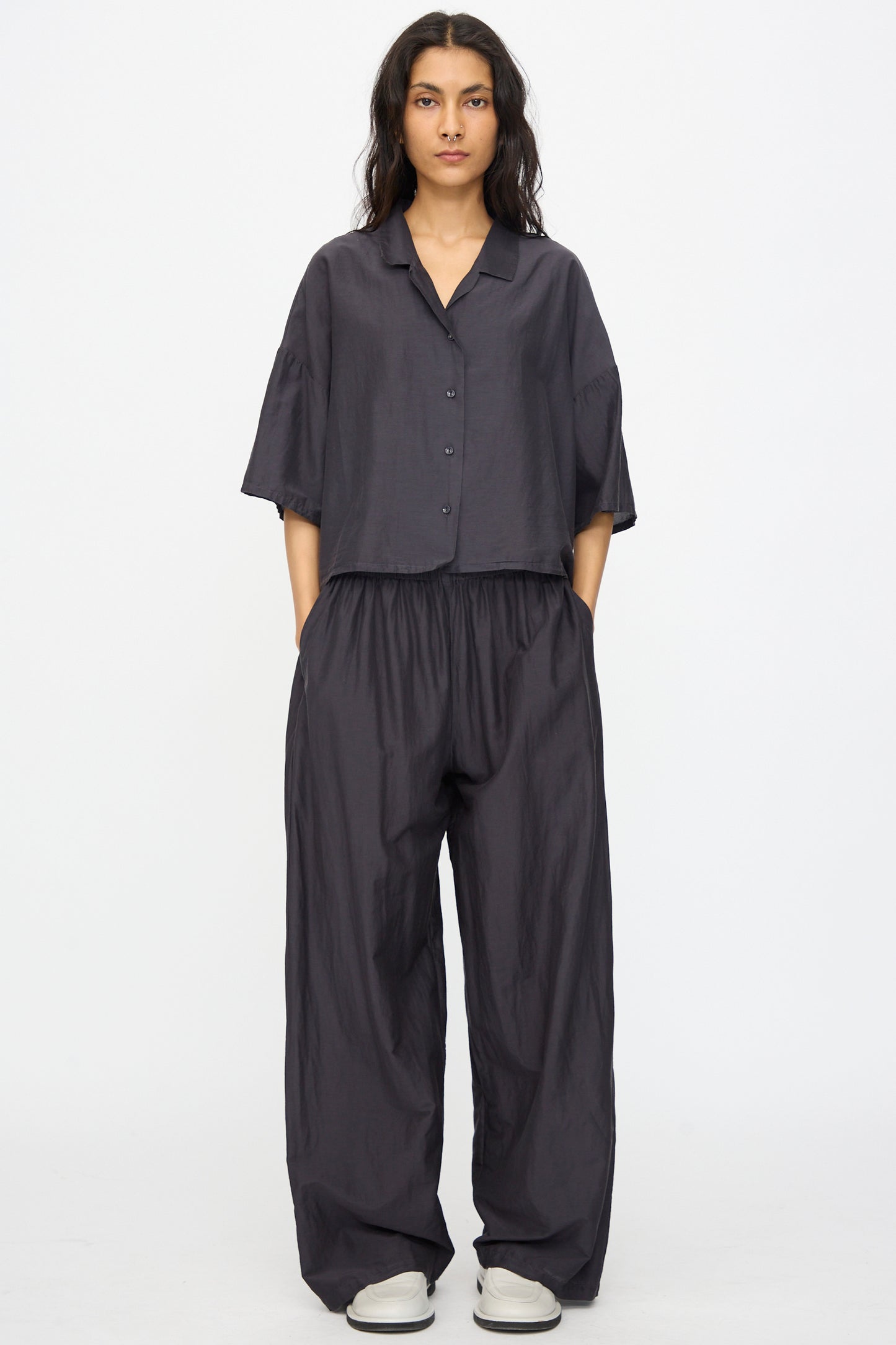 A person stands against a plain background wearing the Black Crane Cropped Shirt in Faded Black with matching wide-leg pants, hands in pockets, and light-colored shoes.