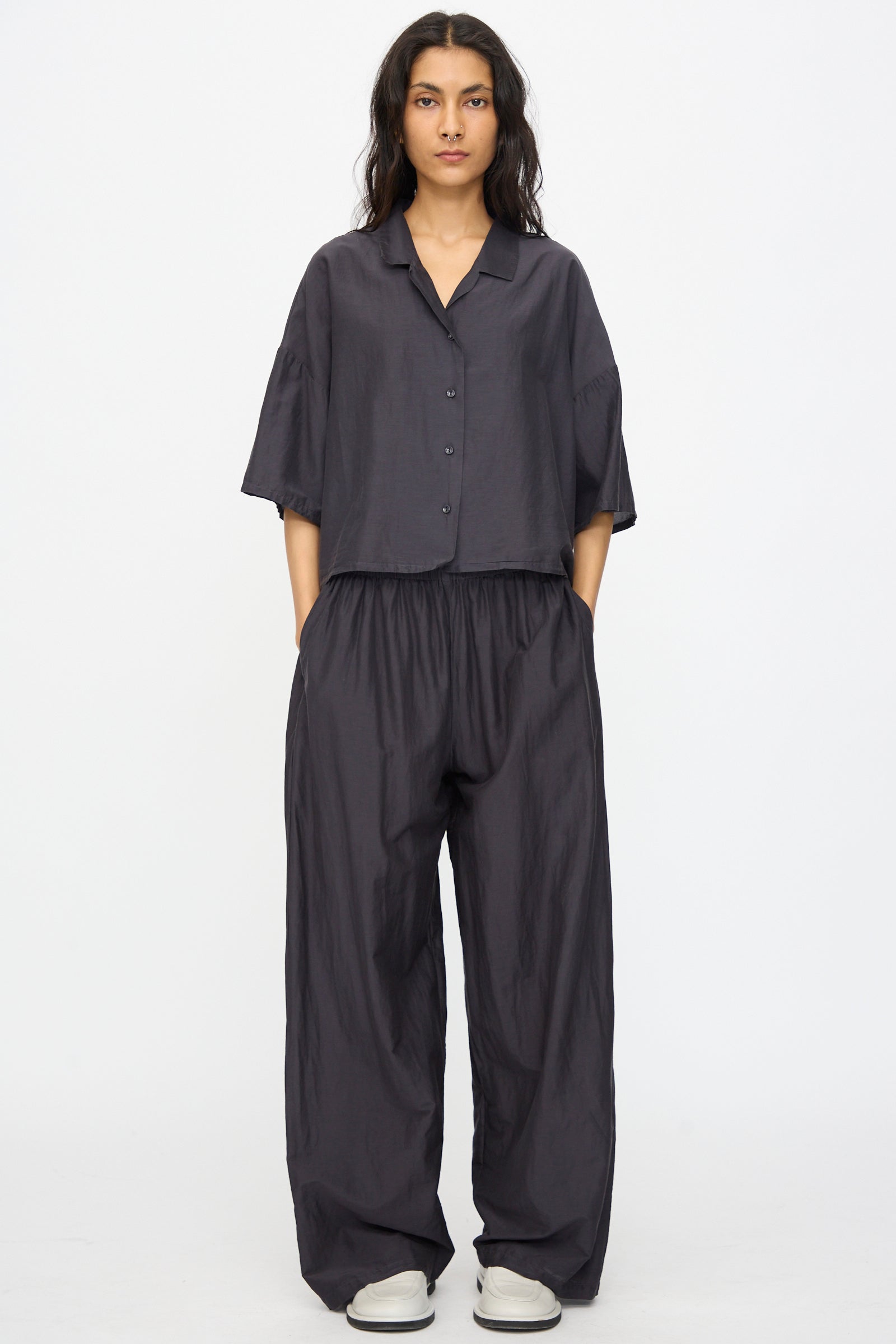 A person stands against a plain background wearing the Black Crane Cropped Shirt in Faded Black with matching wide-leg pants, hands in pockets, and light-colored shoes.