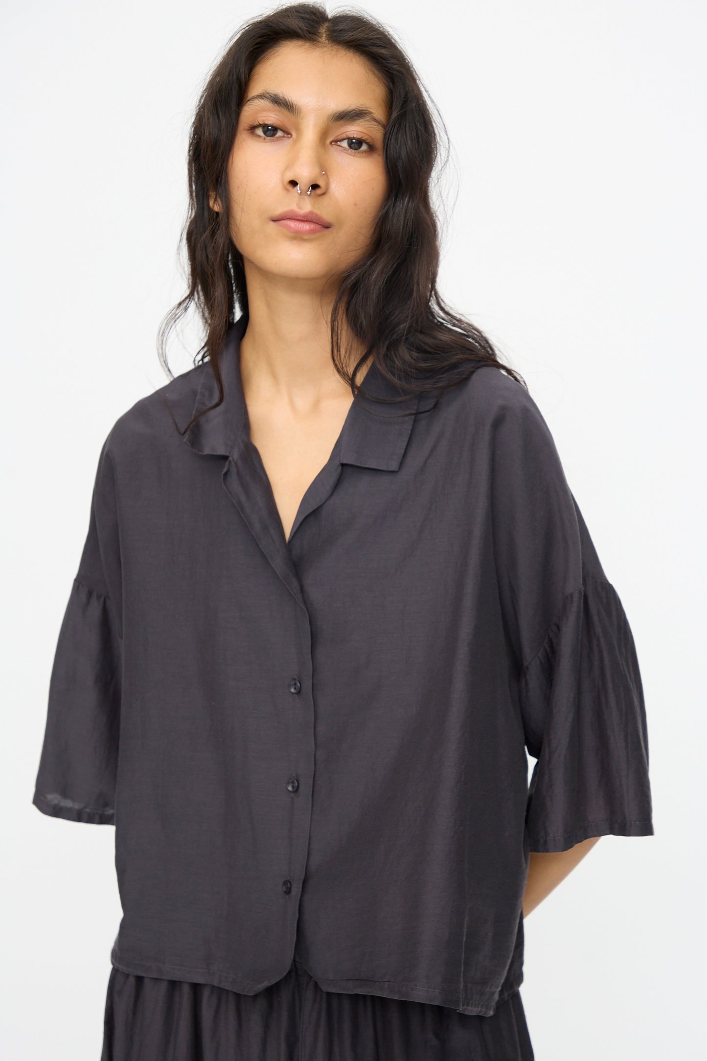Woman with long dark hair wears the Black Crane Cropped Shirt in Faded Black, a boxy fit short sleeve style, standing against a plain light background.
