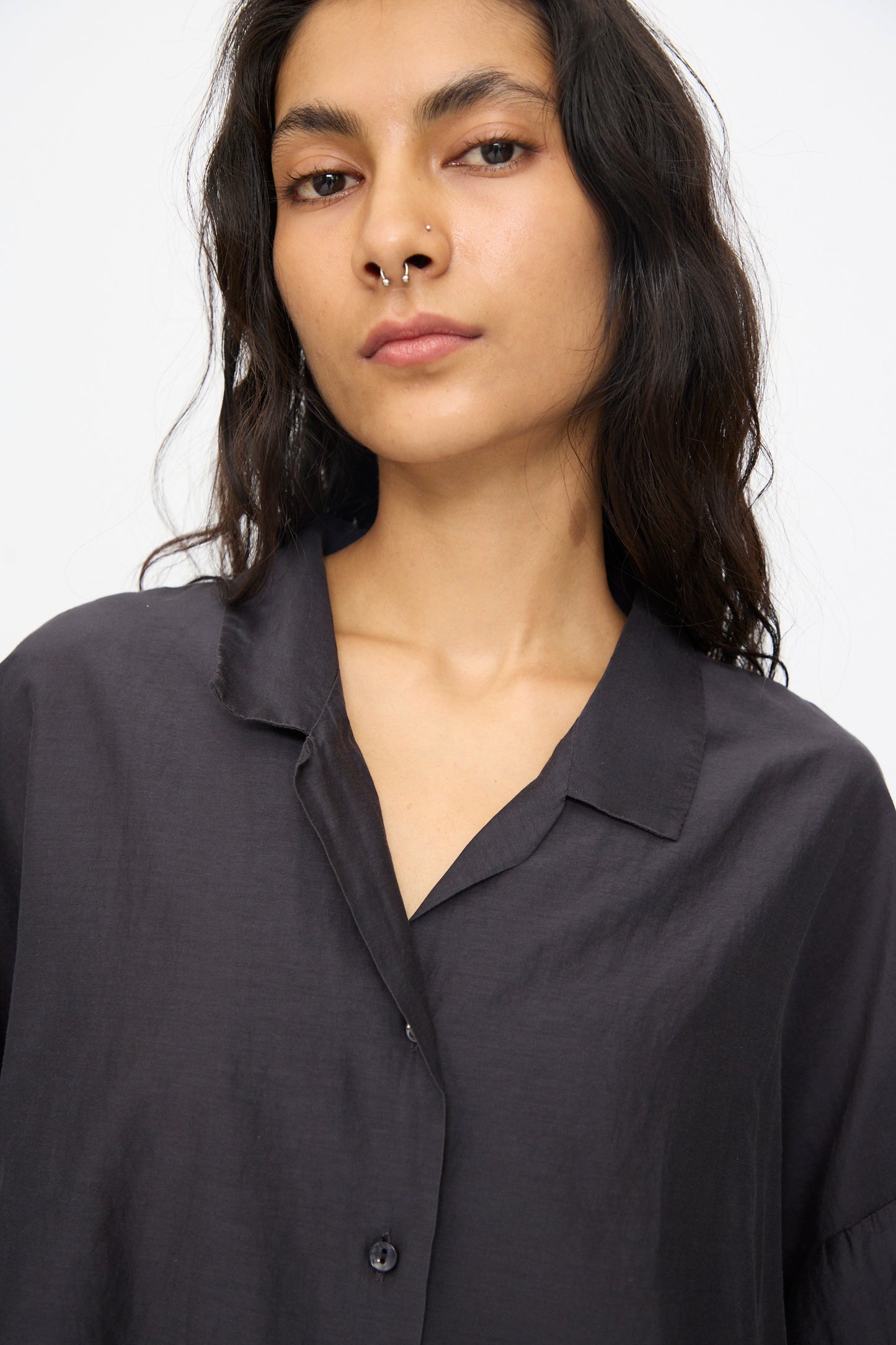 A person with long dark hair and a nose ring models the Black Crane Cropped Shirt in Faded Black, featuring a boxy fit and short sleeves, while looking directly at the camera against a plain light background.