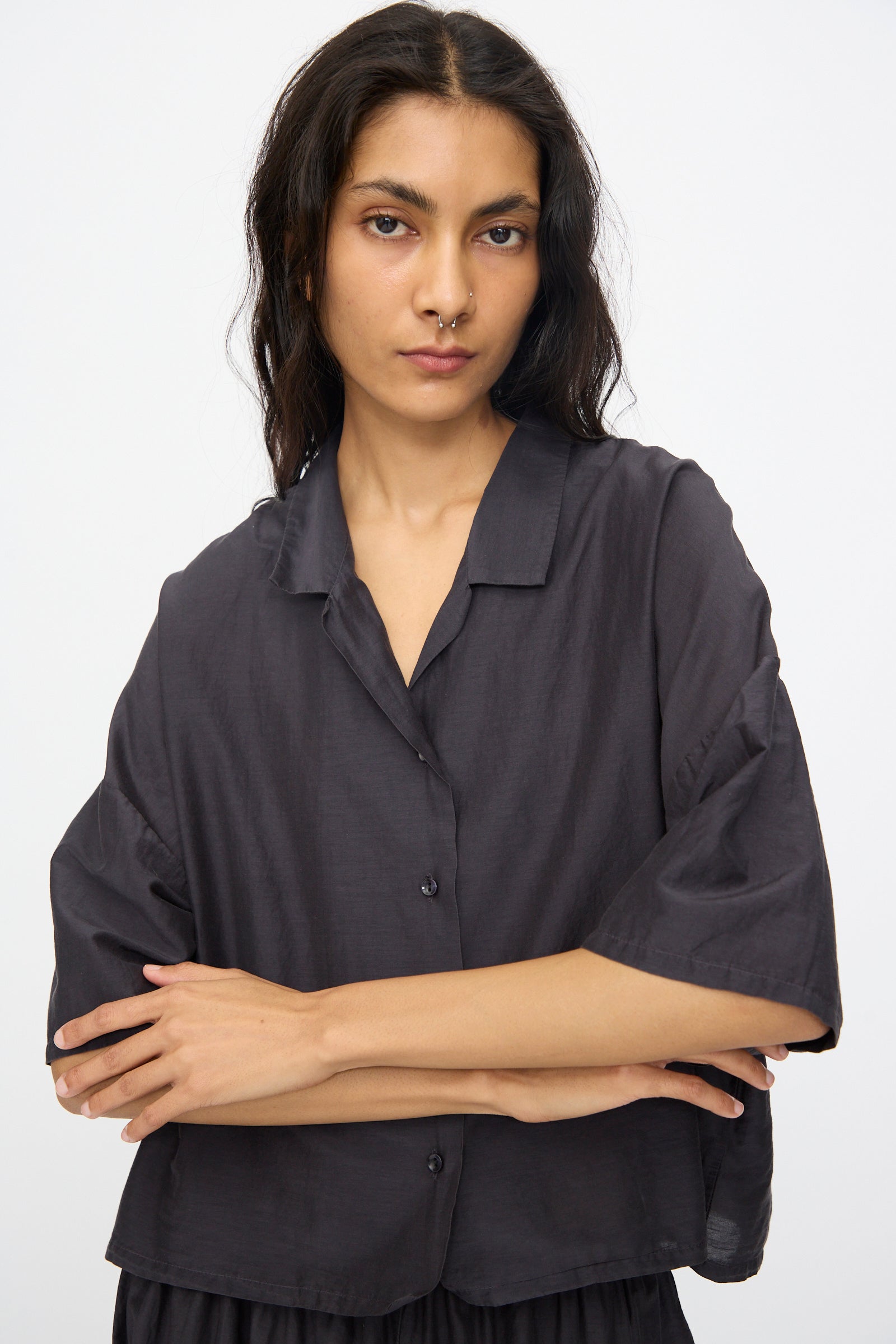 A woman with long dark hair stands with her arms crossed against a plain light background, wearing the Black Crane Cropped Shirt in Faded Black.