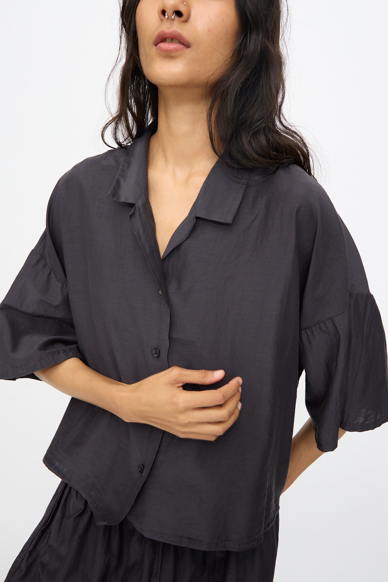 A person wears the Black Crane Cropped Shirt in Faded Black—featuring a loose, boxy fit and cropped short sleeves—as they stand against a plain background, one hand resting on their torso.