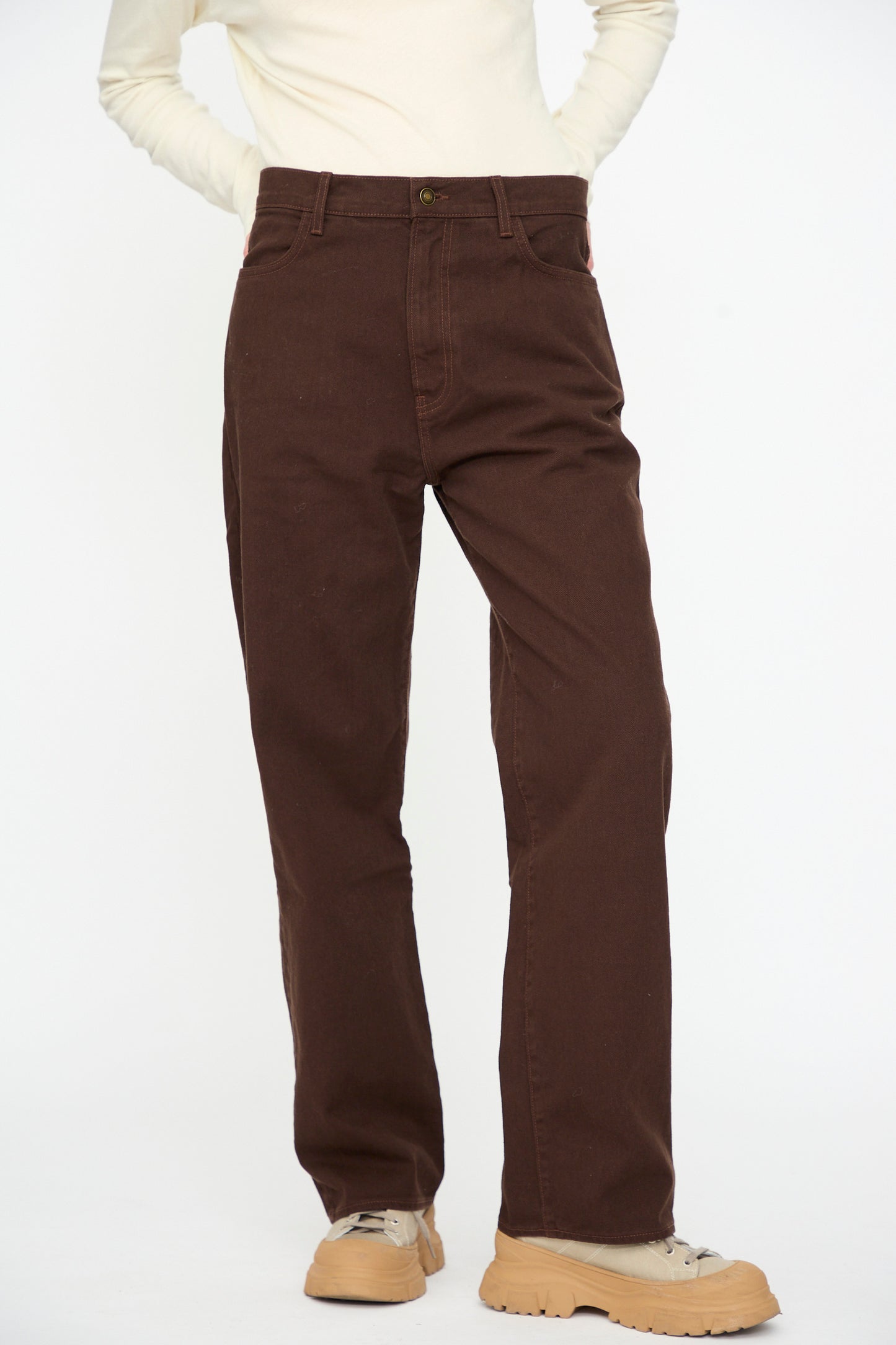 A person wearing the Black Crane Denim Pant in Dark Cacao, paired with an off-white long-sleeve top and beige chunky sneakers, stands against a plain white background.
