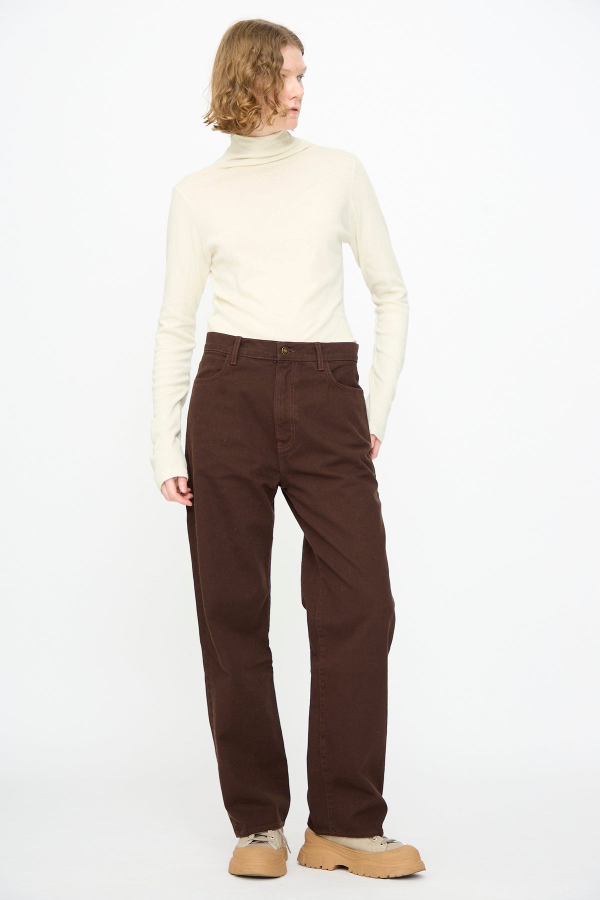 A person wearing a cream turtleneck, Black Crane Denim Pant in Dark Cacao, and tan shoes stands against a plain white background, looking to the side.