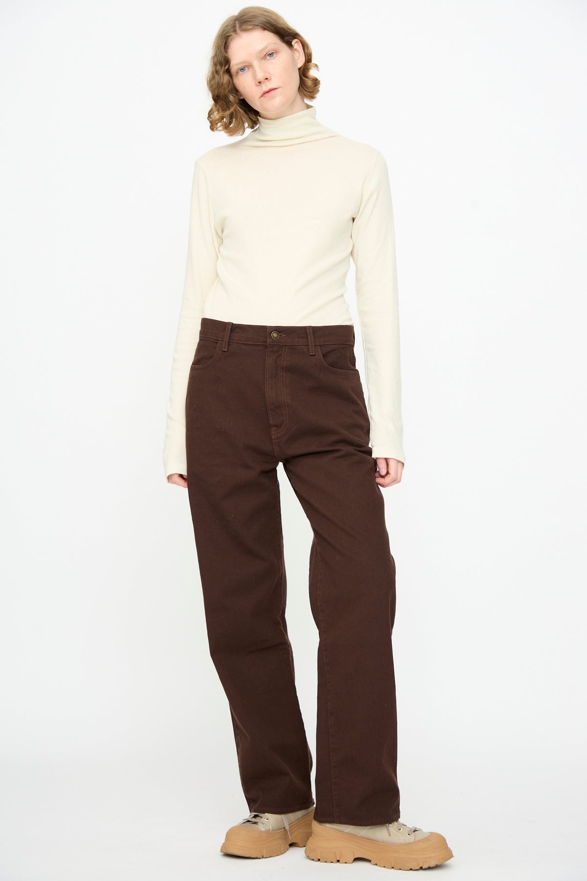A person stands against a white background, wearing a cream turtleneck, Denim Pant in Dark Cacao by Black Crane, and tan shoes.