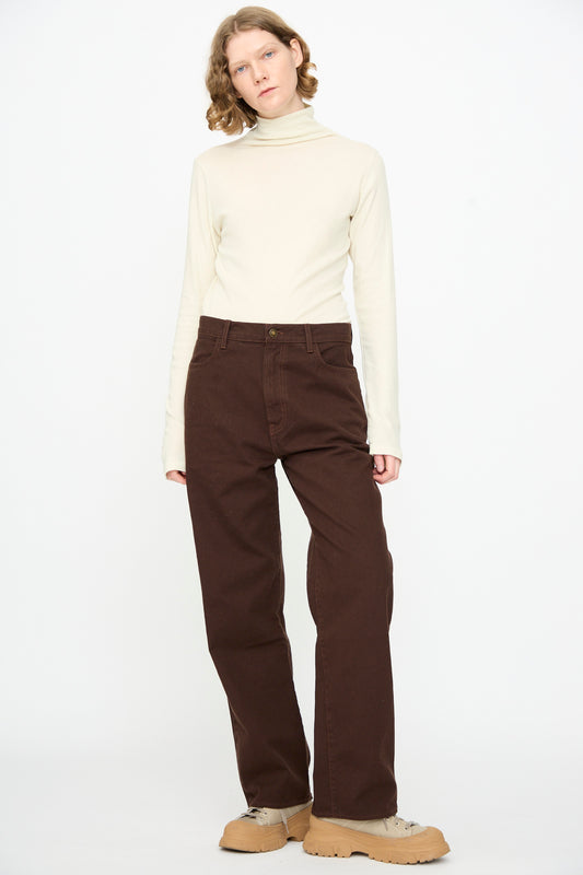 A person stands against a white background, wearing a cream turtleneck, Denim Pant in Dark Cacao by Black Crane, and tan shoes.
