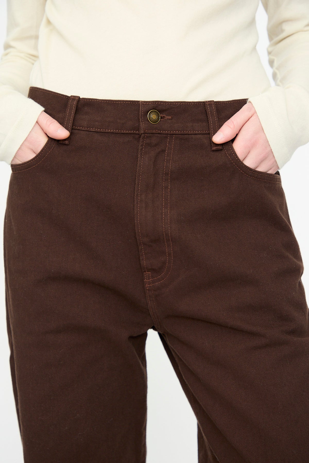A person wearing a cream long-sleeve shirt and Black Crane’s Denim Pant in Dark Cacao, with hands in the front pockets, shown from waist to mid-thigh.