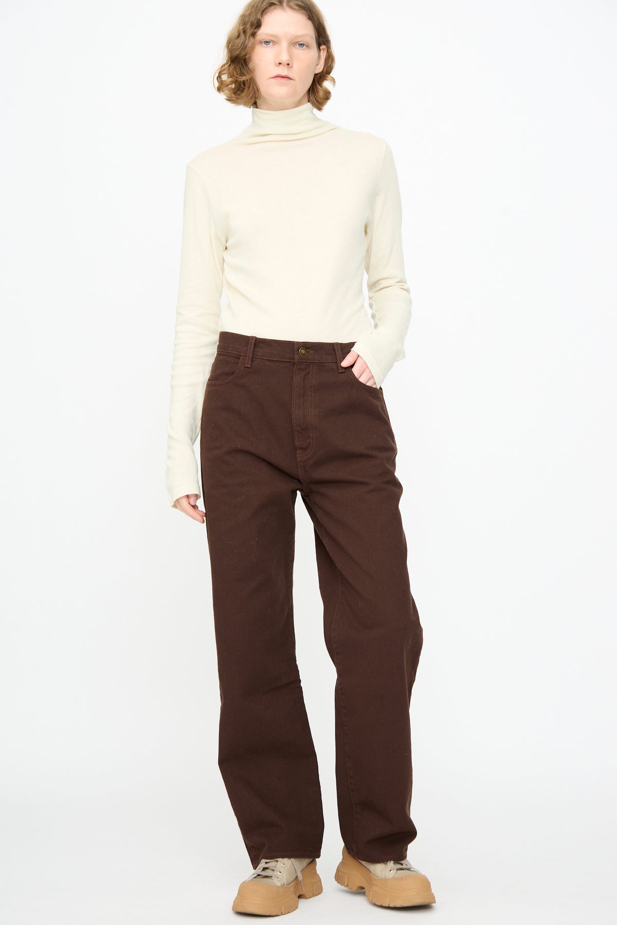 A person stands against a plain background, wearing a cream turtleneck, the Black Crane Denim Pant in Dark Cacao, and tan shoes, with one hand in their pocket.