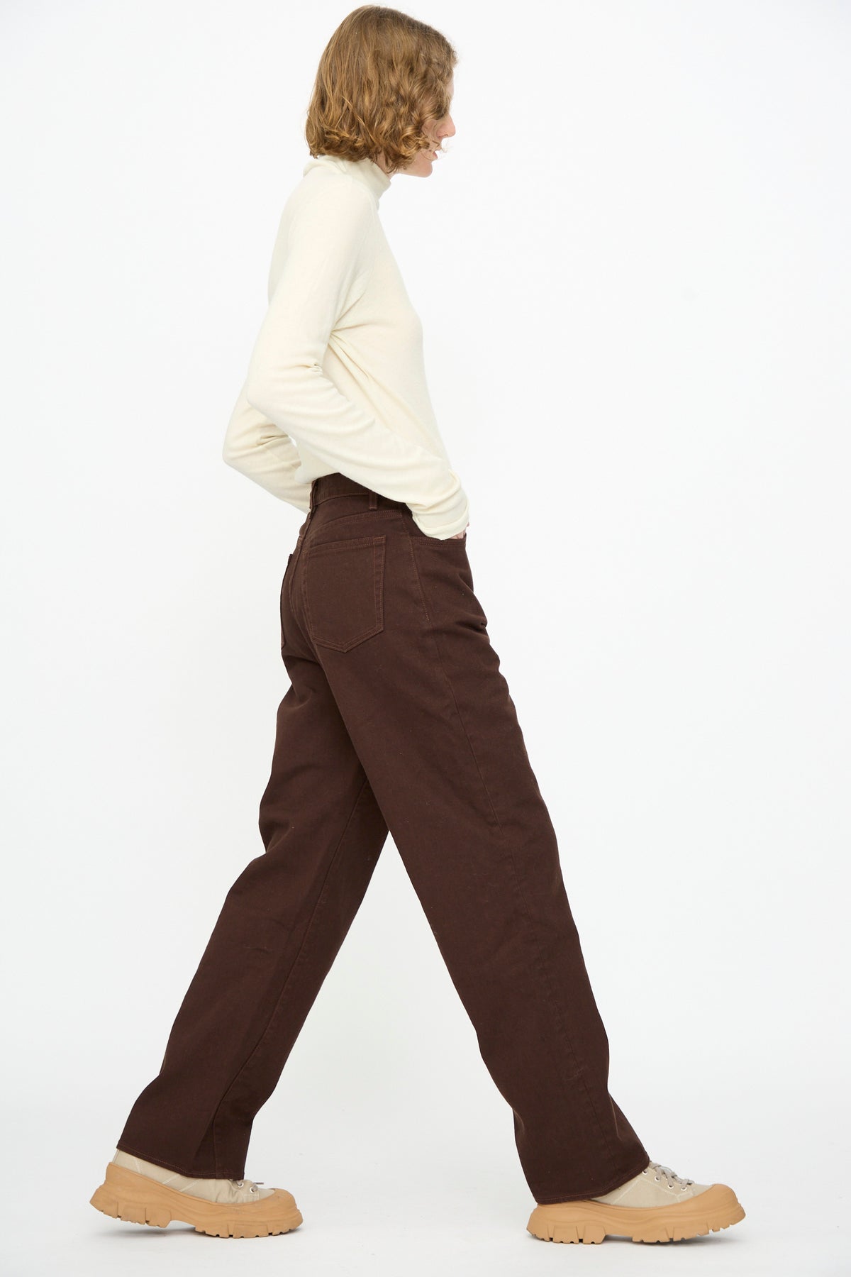 A person stands sideways against a plain white background, wearing a cream long-sleeve top, tan chunky shoes, and the Black Crane Denim Pant in Dark Cacao.