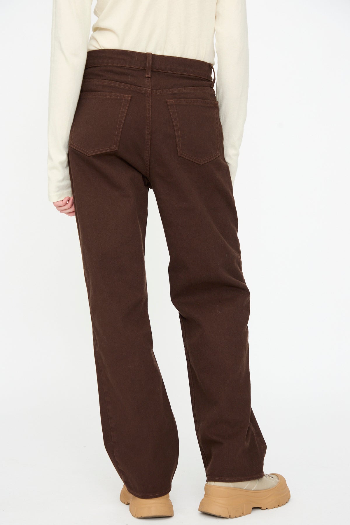 A person wearing a cream long-sleeve top, Black Crane Denim Pant in Dark Cacao, and tan shoes is photographed from the back against a plain white background.