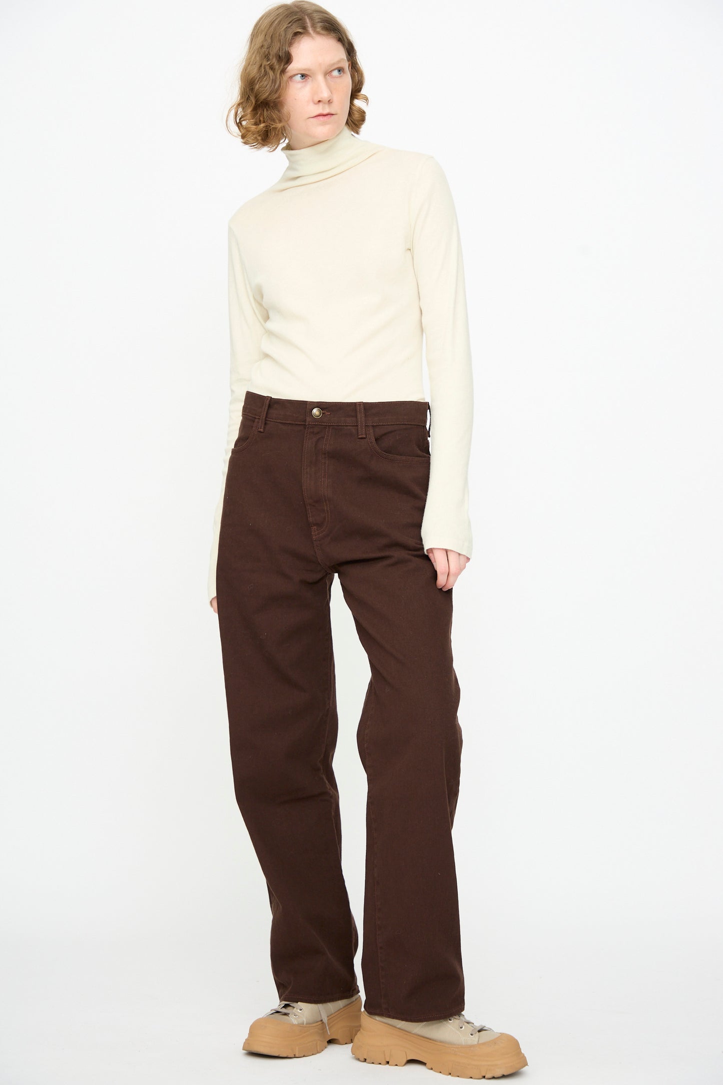 A person stands against a white background, wearing a cream turtleneck, the Black Crane Denim Pant in Dark Cacao, and beige chunky shoes.