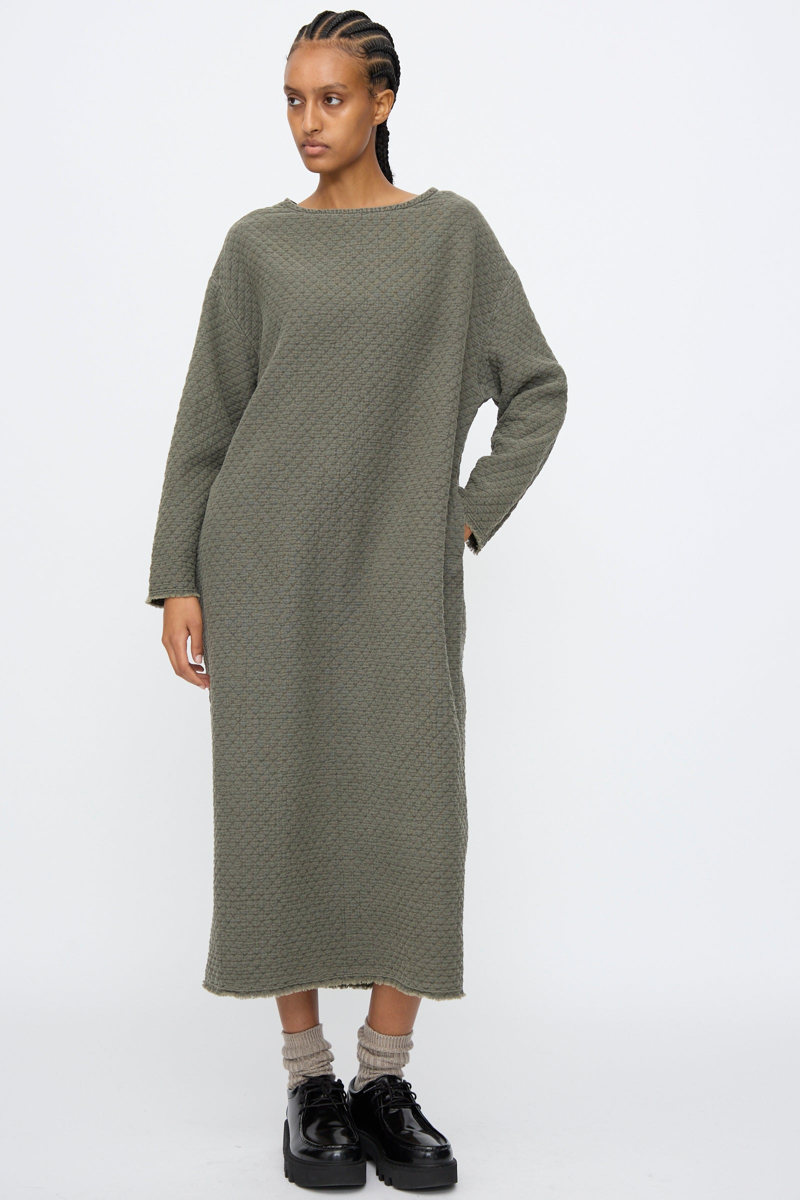 A woman stands against a plain background wearing the Black Crane Diamond Quilted Dress in Slate, a relaxed fit, long-sleeve, ankle-length cotton dress, paired with beige socks and black platform shoes.