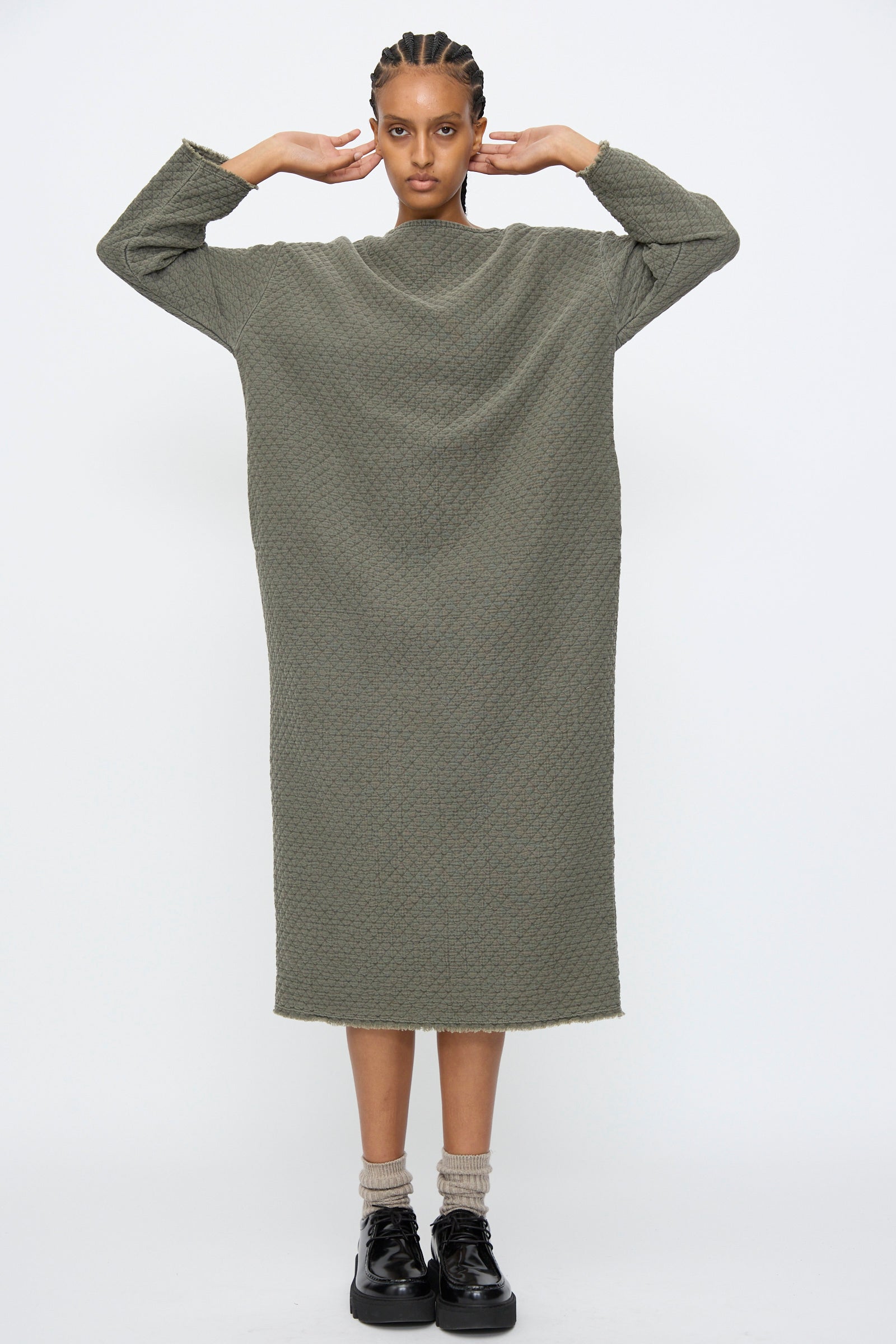 A person stands facing forward in the Black Crane Diamond Quilted Dress in Slate, paired with beige socks and black shoes against a plain white background.