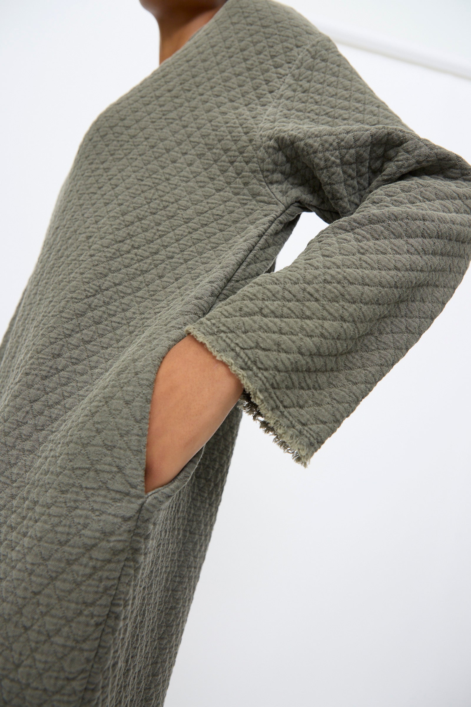 A person wearing the Black Crane Diamond Quilted Dress in Slate, featuring textured quilted cotton, long sleeves, a relaxed fit, and a hand in the side pocket.