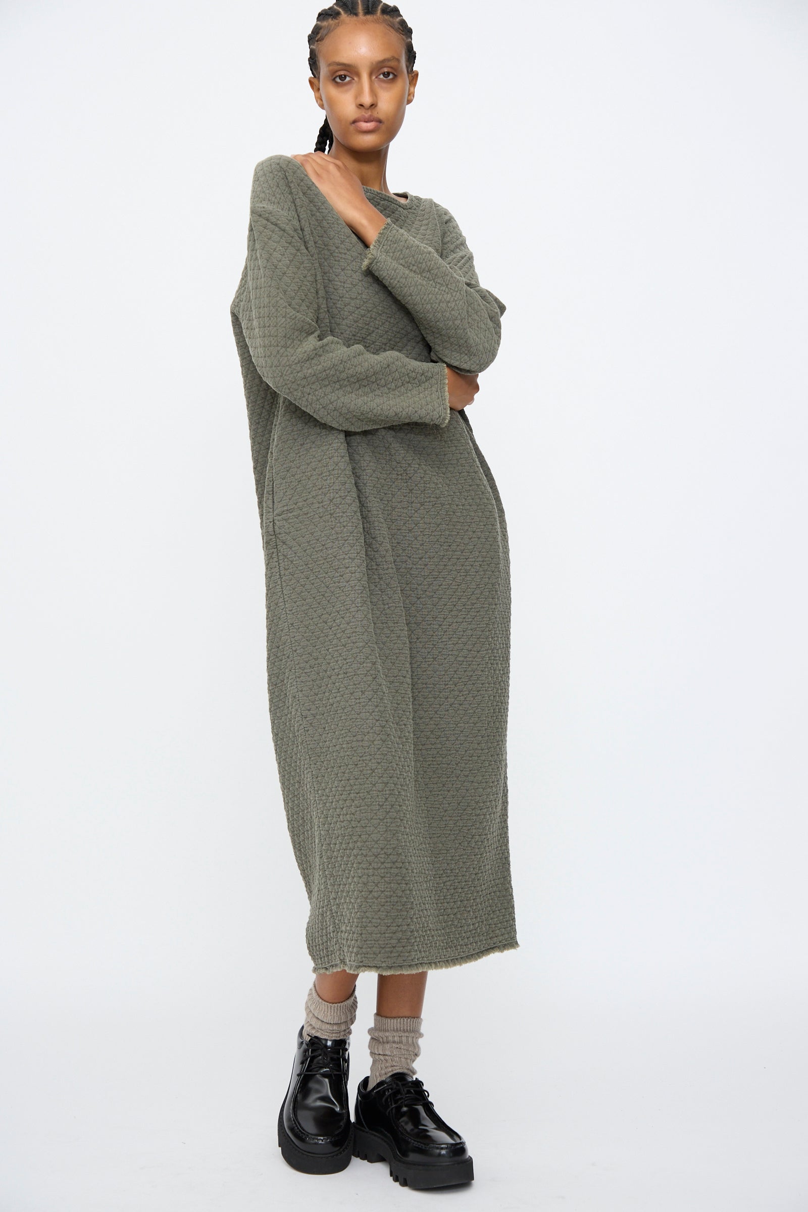 A person stands against a plain background wearing the Black Crane Diamond Quilted Dress in Slate, styled with beige socks and black platform shoes.