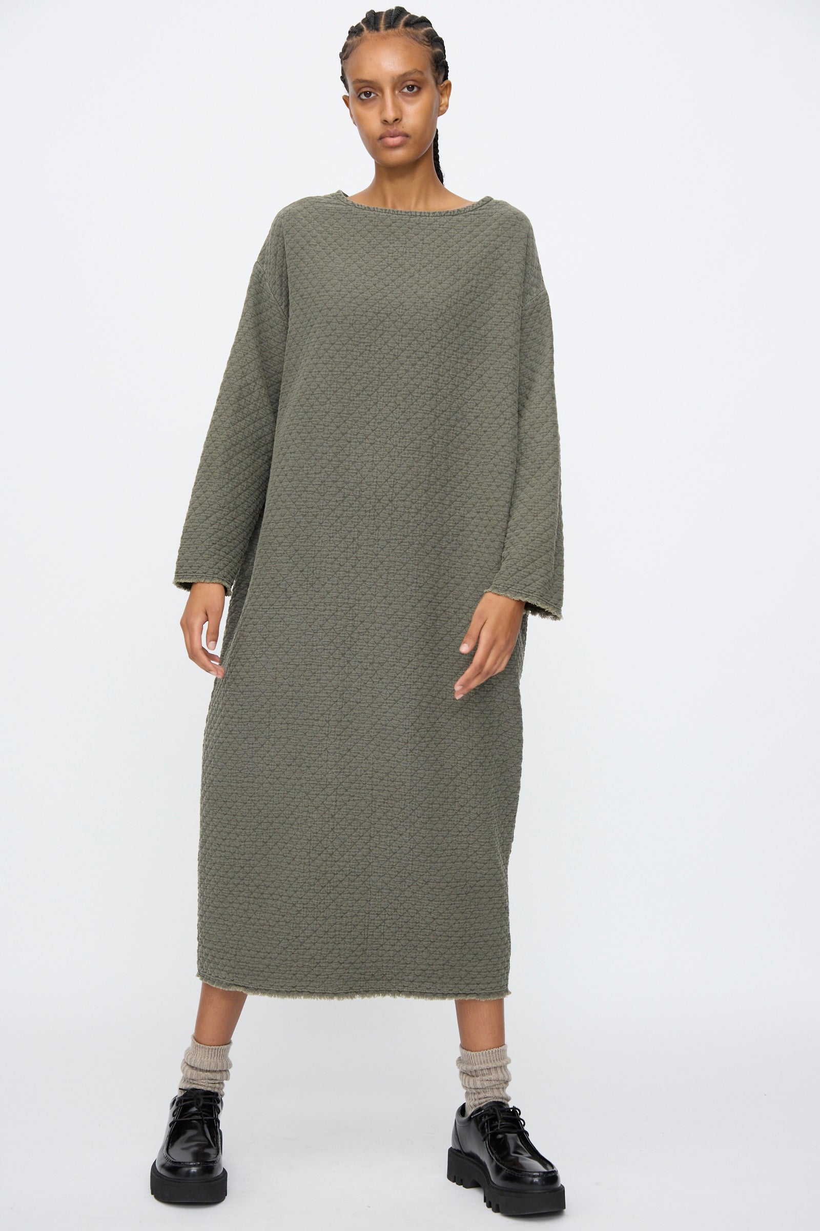A person wears the Black Crane Diamond Quilted Dress in Slate, paired with beige socks and black chunky shoes, standing against a plain white background.