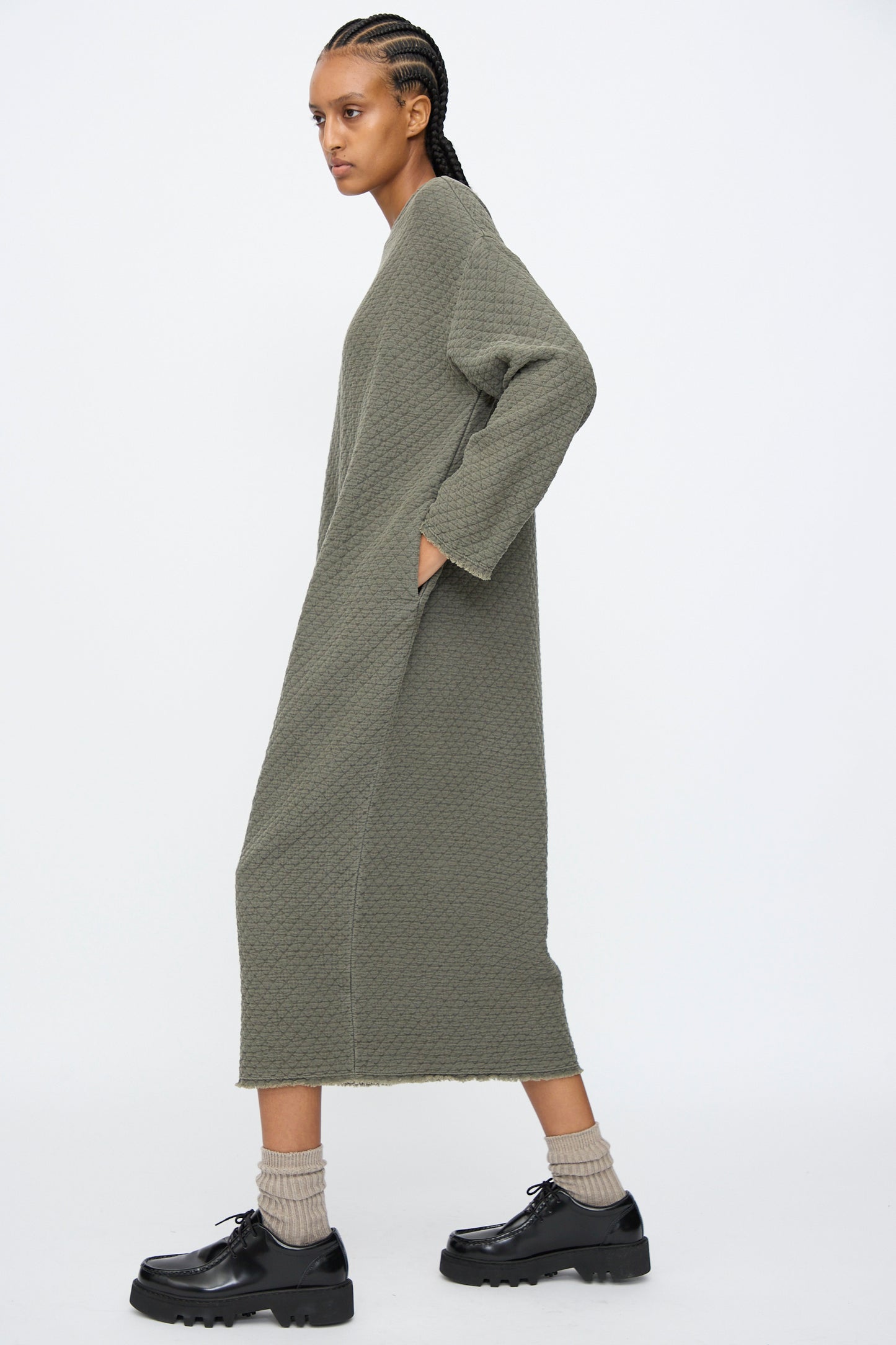 A person stands sideways wearing the Black Crane Diamond Quilted Dress in Slate, styled with black chunky shoes, beige socks, and braided hair.