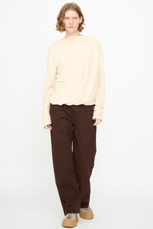 A person with shoulder-length hair stands against a white background, wearing the Black Crane Double Layer Draped Top in Natural, brown wide-leg pants, and tan sneakers.