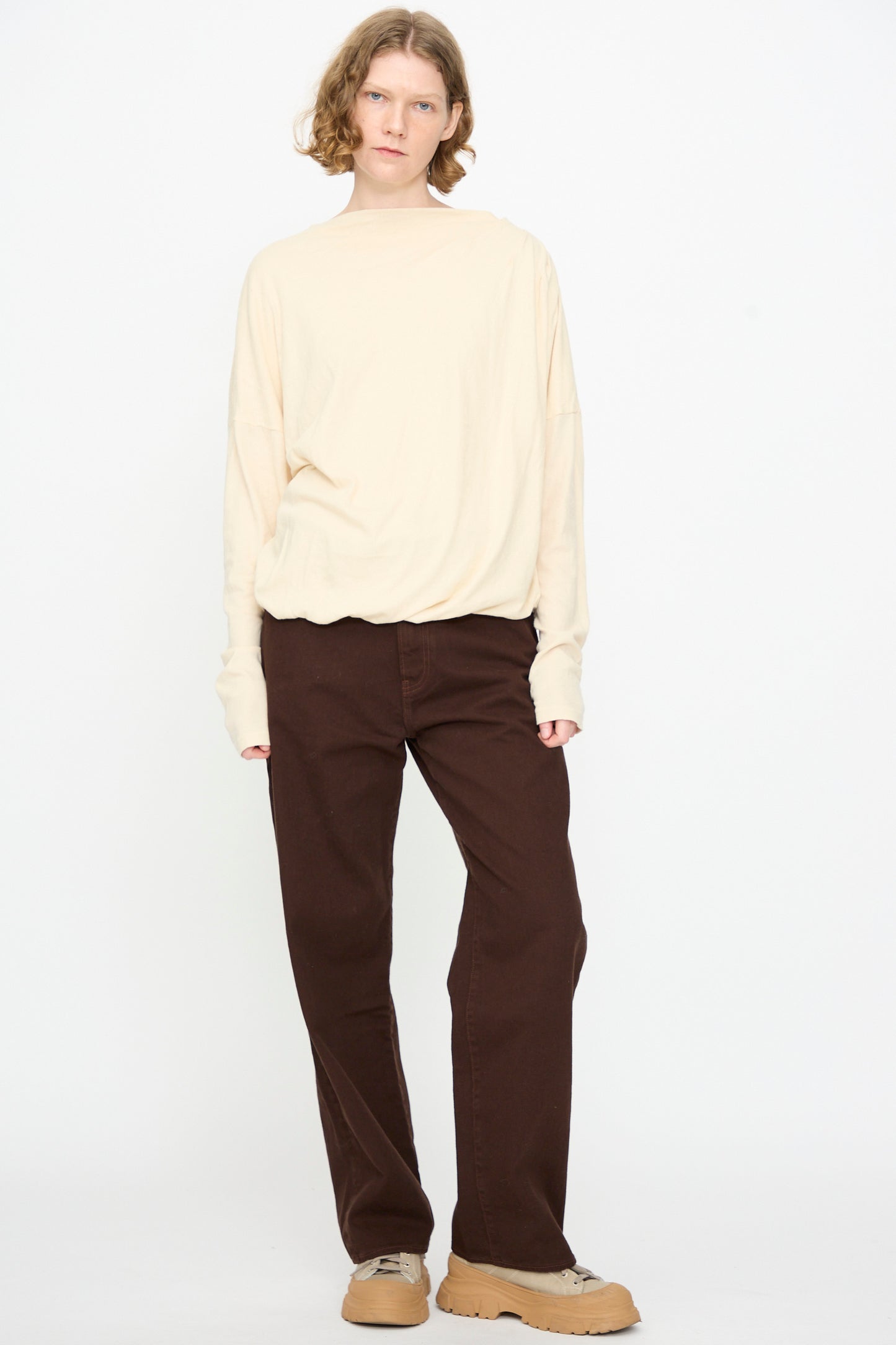 Person standing against a white background, wearing the Black Crane Double Layer Draped Top in Natural, paired with dark brown pants and tan sneakers.