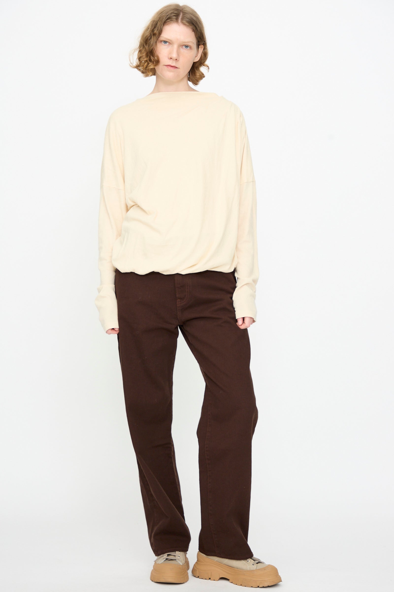 Person standing against a white background, wearing the Black Crane Double Layer Draped Top in Natural, paired with dark brown pants and tan sneakers.