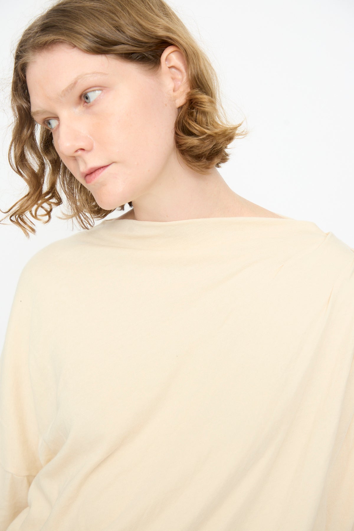 A person with short, wavy, light brown hair wears the Black Crane Double Layer Draped Top in Natural and looks slightly to the side against a plain white background.
