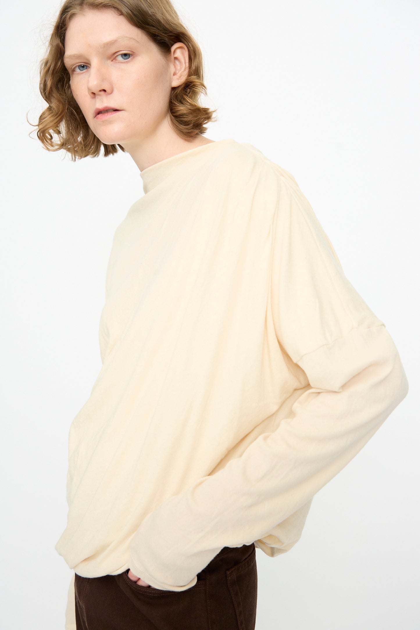 A person with short, wavy hair models the Black Crane Double Layer Draped Top in Natural—a relaxed fit, off-white cotton cashmere long sleeve shirt—paired with brown pants, posing against a plain white background.