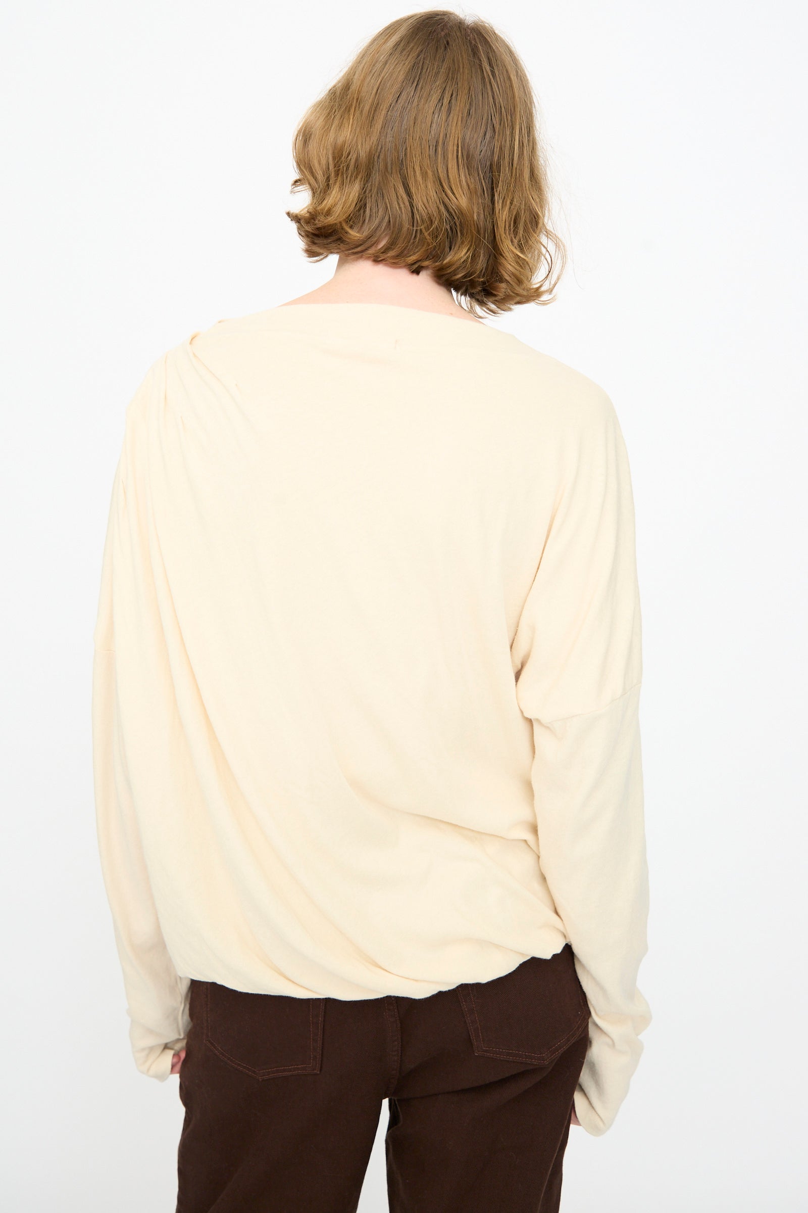 A person with shoulder-length light brown hair, wearing the Black Crane Double Layer Draped Top in Natural and brown pants, is standing with their back to the camera against a plain white background.