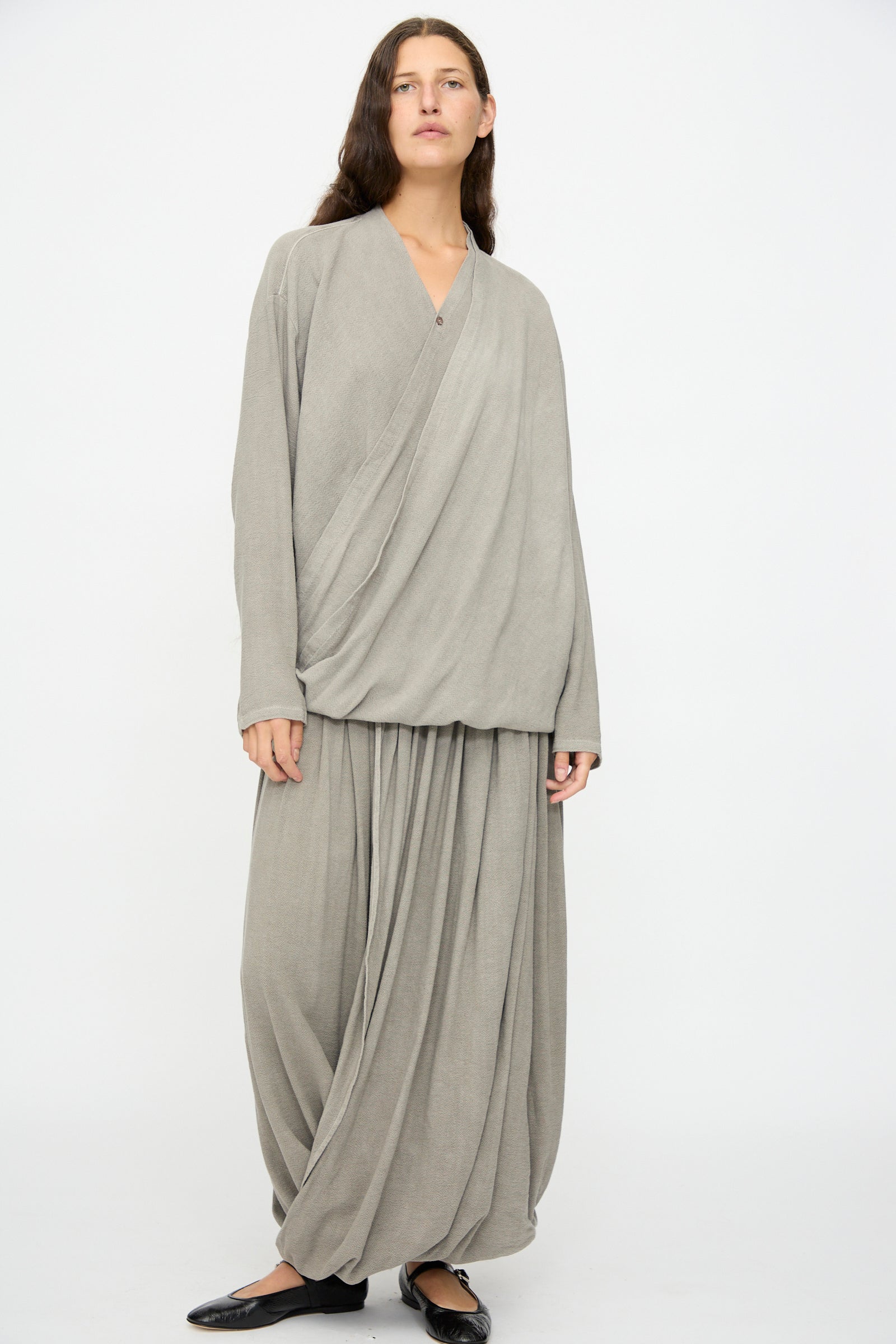 A person stands against a plain background wearing Black Crane's Double Layered Drapy Skirt in Dark Stone—a loose, cotton rayon blend midi with long sleeves—paired with black flat shoes.