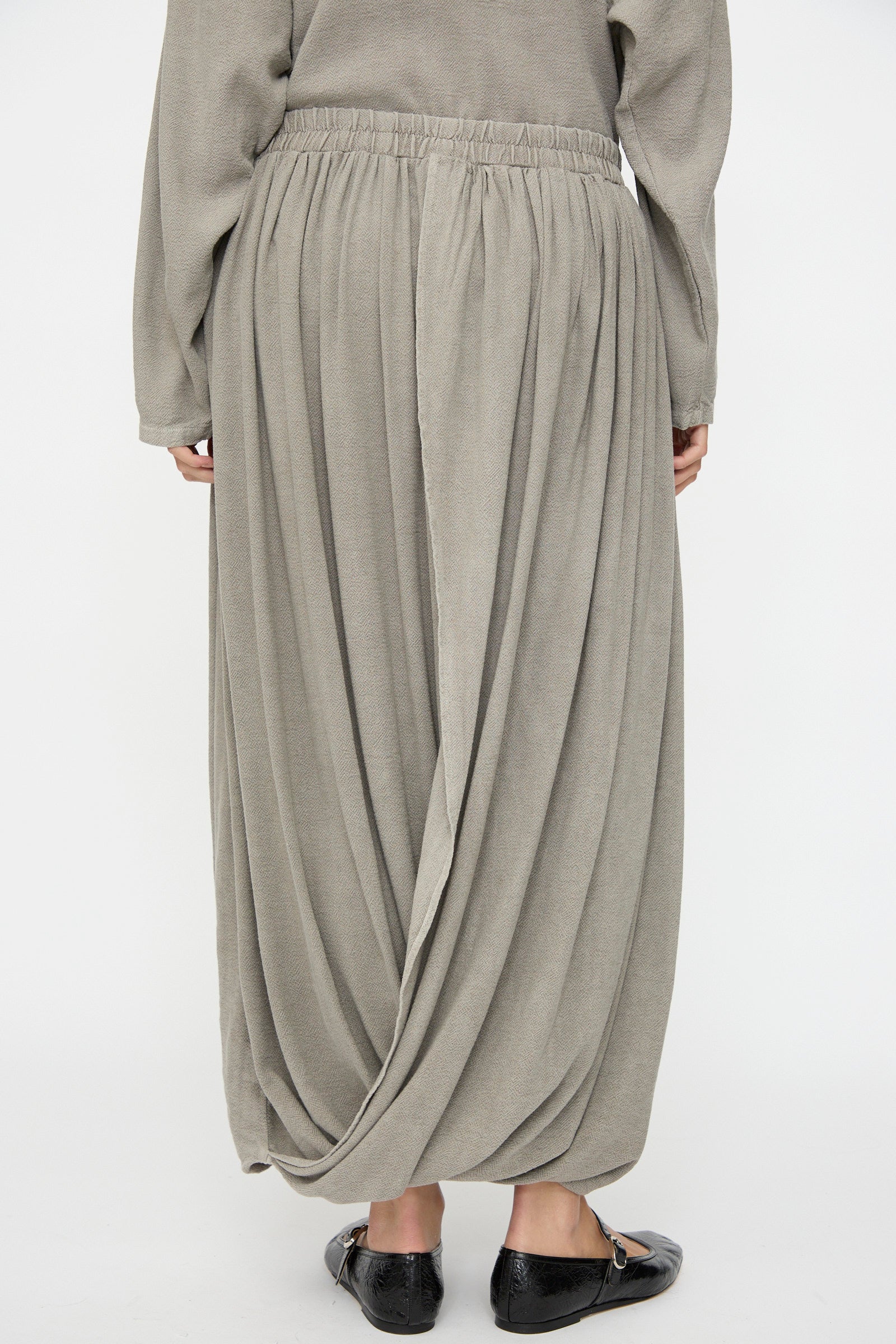 A person is seen from the back wearing the Black Crane Double Layered Drapy Skirt in Dark Stone, a light grey cotton-rayon midi with an elastic waistband, paired with black flat shoes.