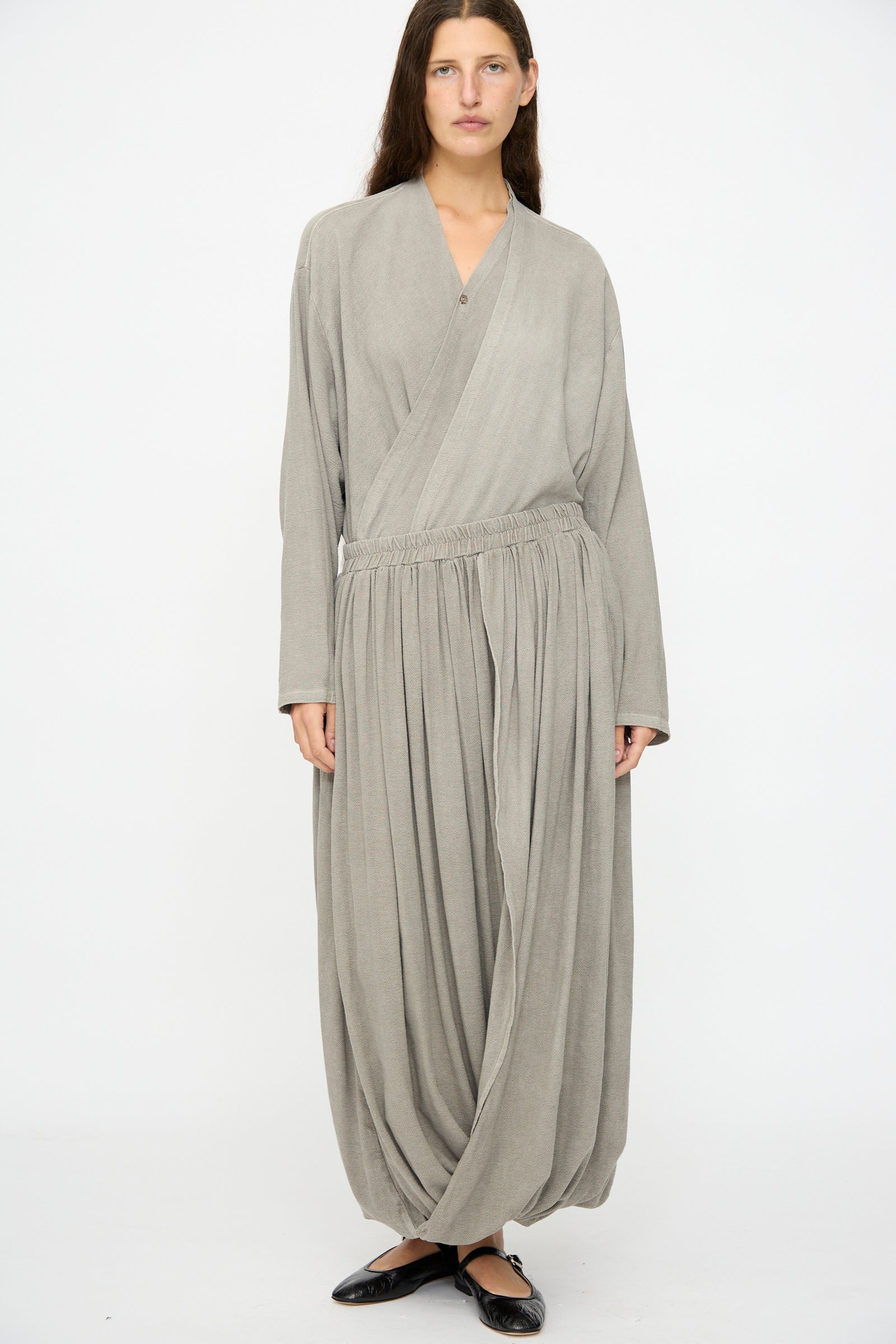 A person stands against a plain background wearing a loose, light gray wrap top paired with Black Crane's Double Layered Drapy Skirt in Dark Stone, featuring an elastic waistband and wide drape, finished with black flat shoes.