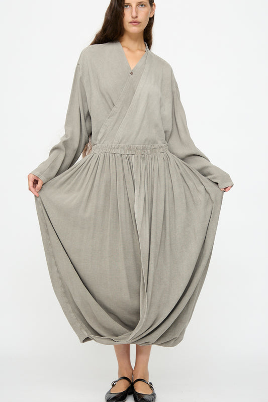 A woman stands against a white background wearing the Black Crane Double Layered Drapy Skirt in Dark Stone—a loose-fitting, midi-length skirt with an elastic waistband—paired with a long-sleeved top.