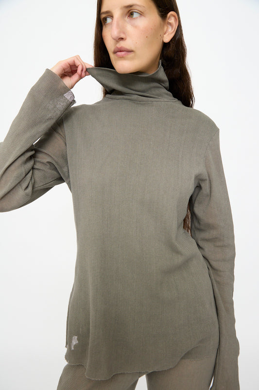 A person models the Black Crane Double Mesh Turtleneck in Dark Stone, pulling the collar away from their neck while gazing off to the side against a plain background.