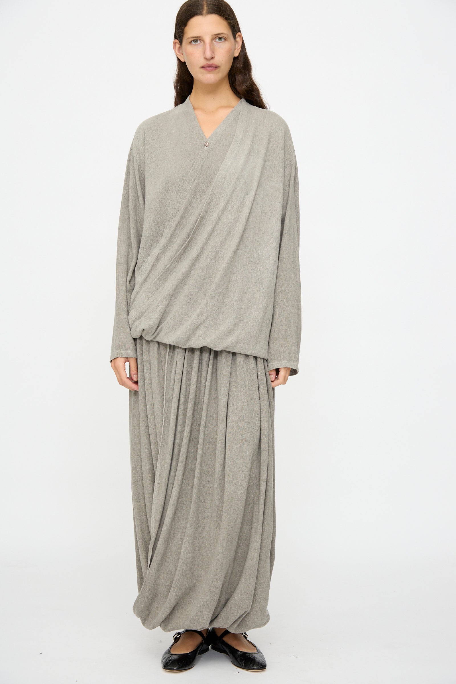A person stands against a plain white background, wearing the Black Crane Drapy Shirt in Dark Stone with a draped front skirt and black flats.