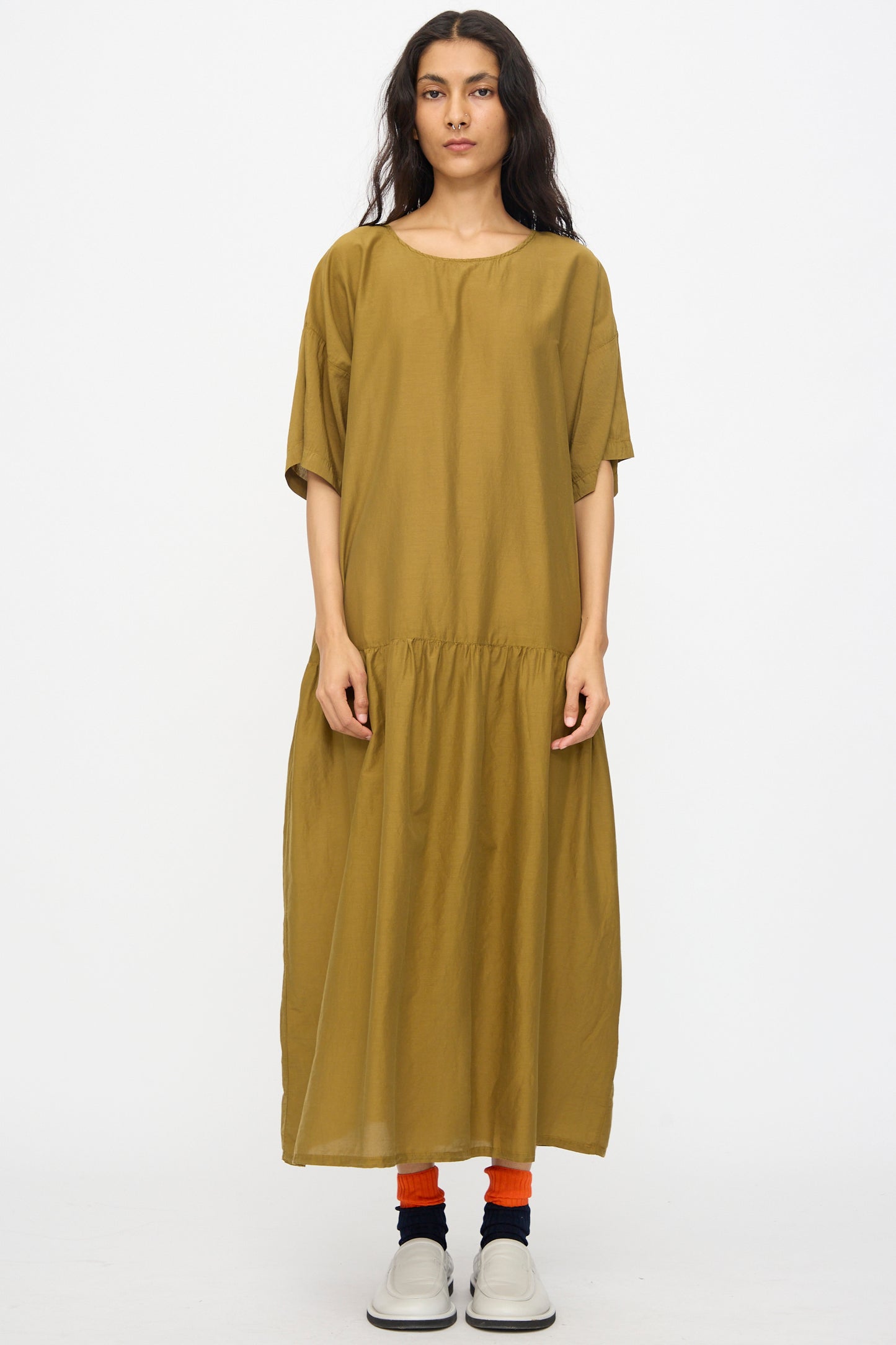 A person stands facing forward in the Black Crane Easy Dress in Dark Olive, paired with bright orange socks and white slip-on shoes, against a plain white background.