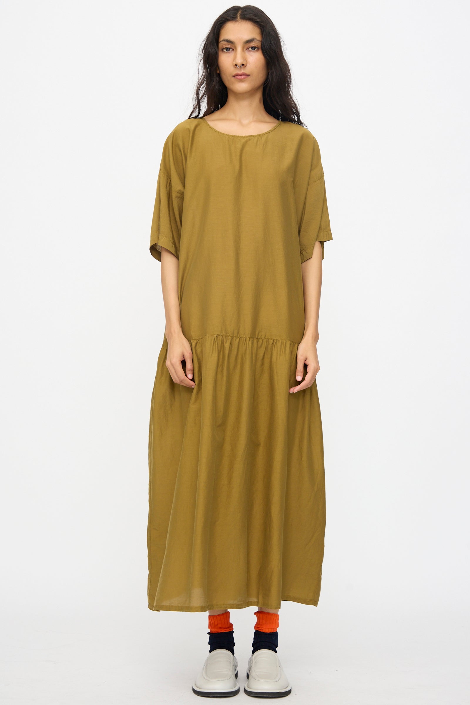 A person stands facing forward in the Black Crane Easy Dress in Dark Olive, paired with bright orange socks and white slip-on shoes, against a plain white background.