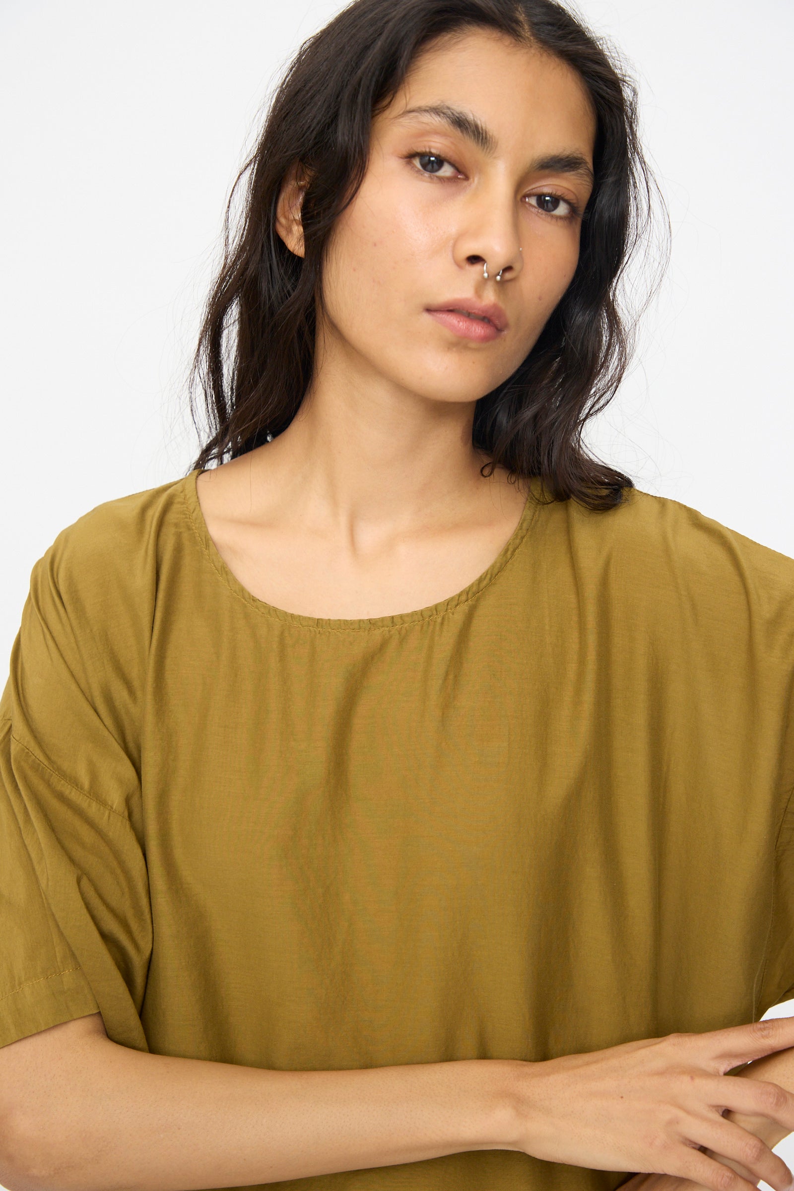 A person with long dark hair and a nose ring wears the Black Crane Easy Dress in Dark Olive, looking directly at the camera against a plain light background.