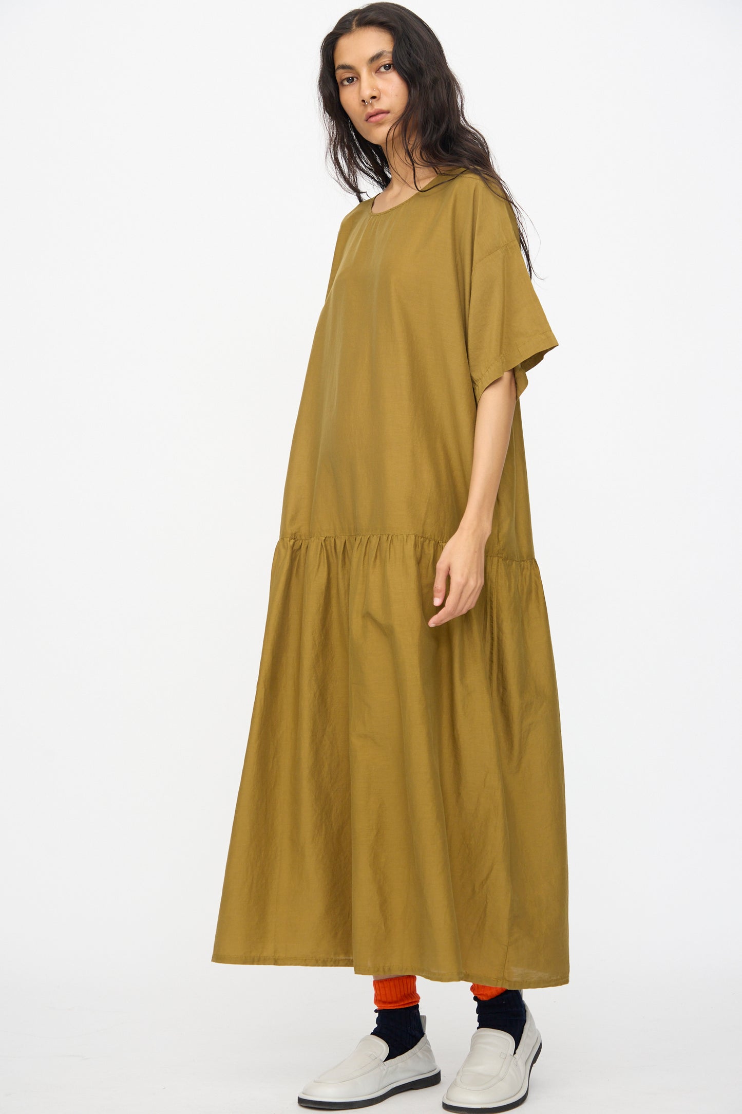 A person stands wearing the Black Crane Easy Dress in Dark Olive, a loose-fitting, short sleeve maxi dress, paired with black socks, red leg warmers, and white slip-on shoes against a white background.