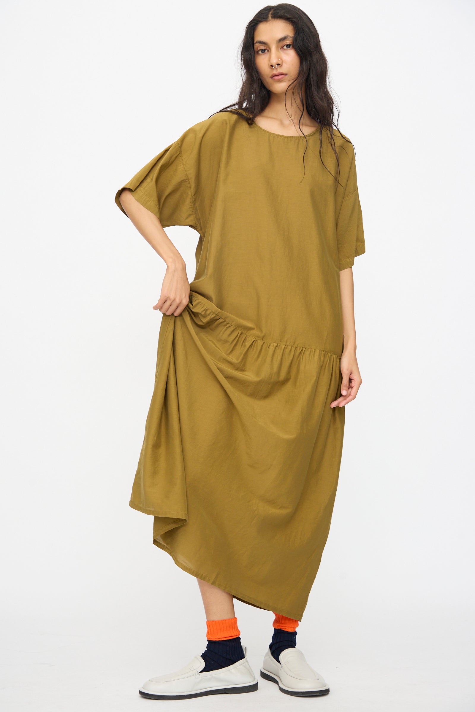 A woman wears the Black Crane Easy Dress in Dark Olive—an oversized, short-sleeve maxi—paired with black socks, orange leg warmers, and white slip-ons, standing against a plain white background.
