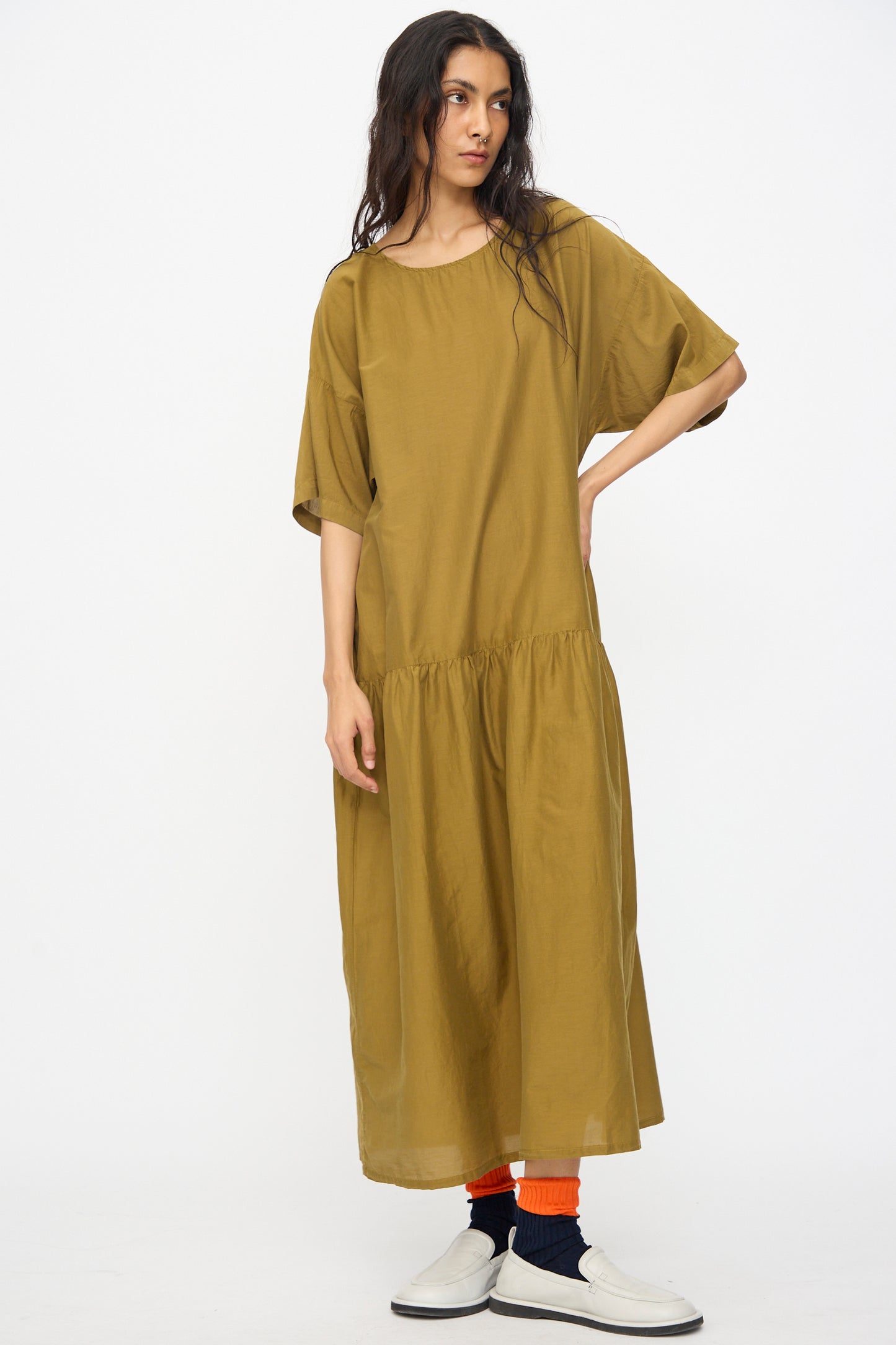 A woman with long hair wears the Black Crane Easy Dress in Dark Olive, an oversized cotton silk midi dress with short sleeves. She stands against a plain white background, pairing white shoes with orange and black socks.