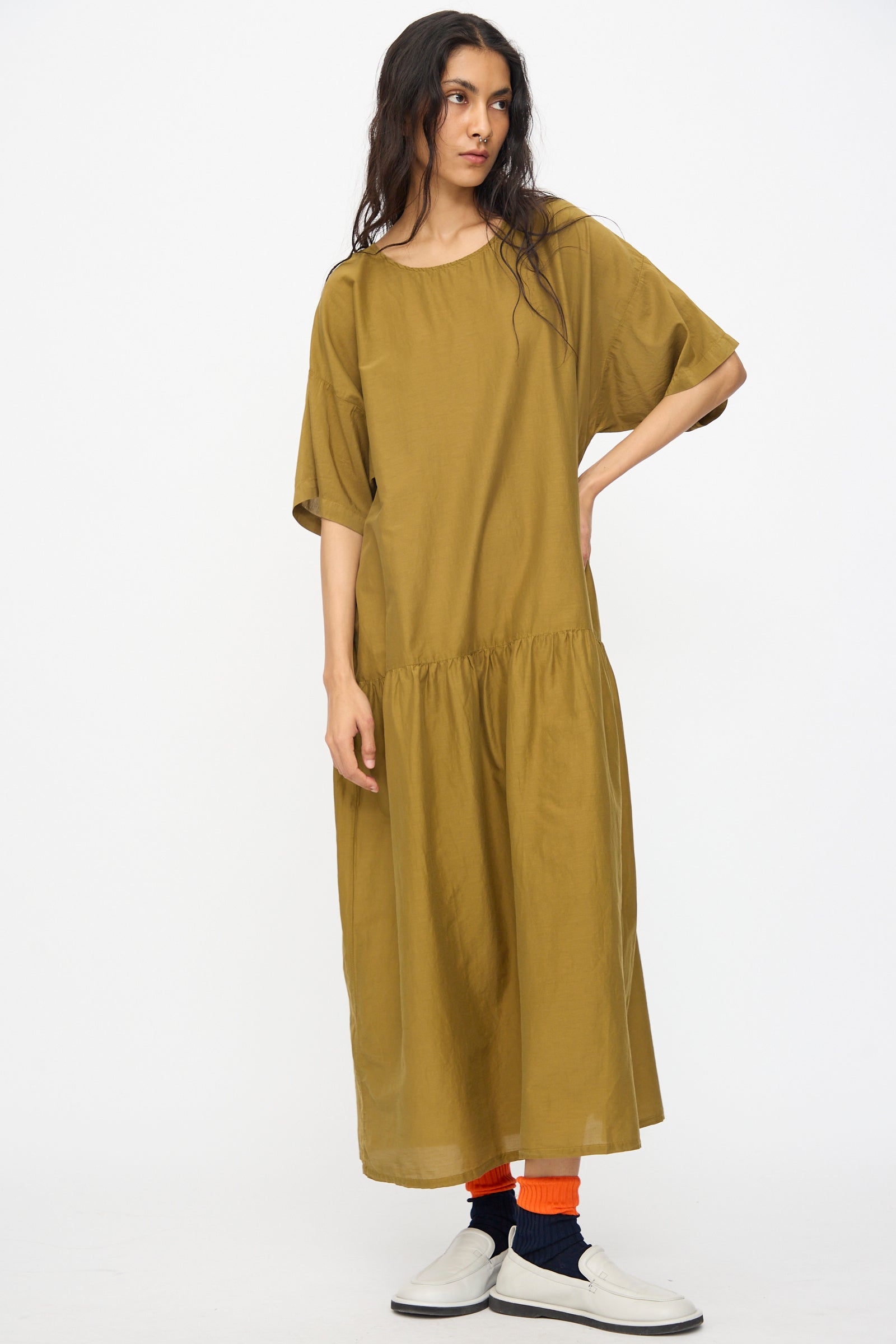 A woman with long hair wears the Black Crane Easy Dress in Dark Olive, an oversized cotton silk midi dress with short sleeves. She stands against a plain white background, pairing white shoes with orange and black socks.