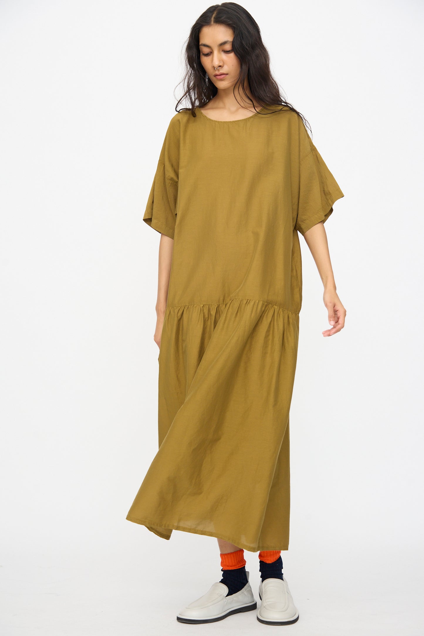 Model wearing the Black Crane Easy Dress in Dark Olive, styled with orange socks and white slip-on shoes, standing against a plain white background.