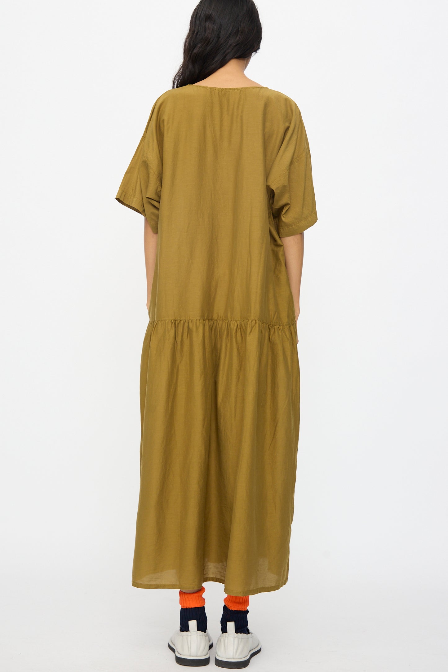 A person stands with their back to the camera wearing the Black Crane Easy Dress in Dark Olive, paired with orange socks and white sneakers, set against a plain white background.