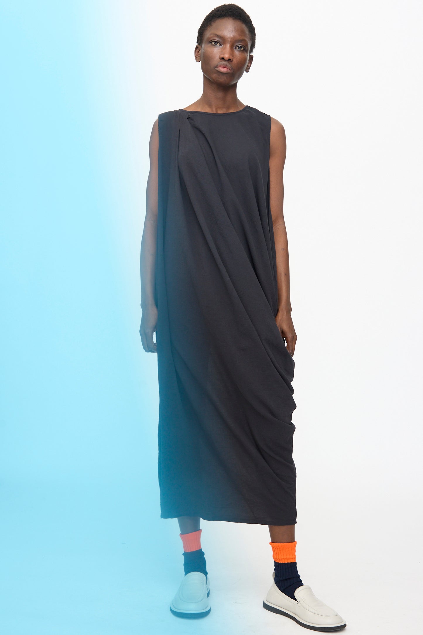 A person stands against a plain background wearing the Black Crane Greek Column Dress in Sumi Black, paired with orange and navy socks and white slip-on shoes. The dress’s relaxed fit flows as a blue gradient overlays the image’s left side.