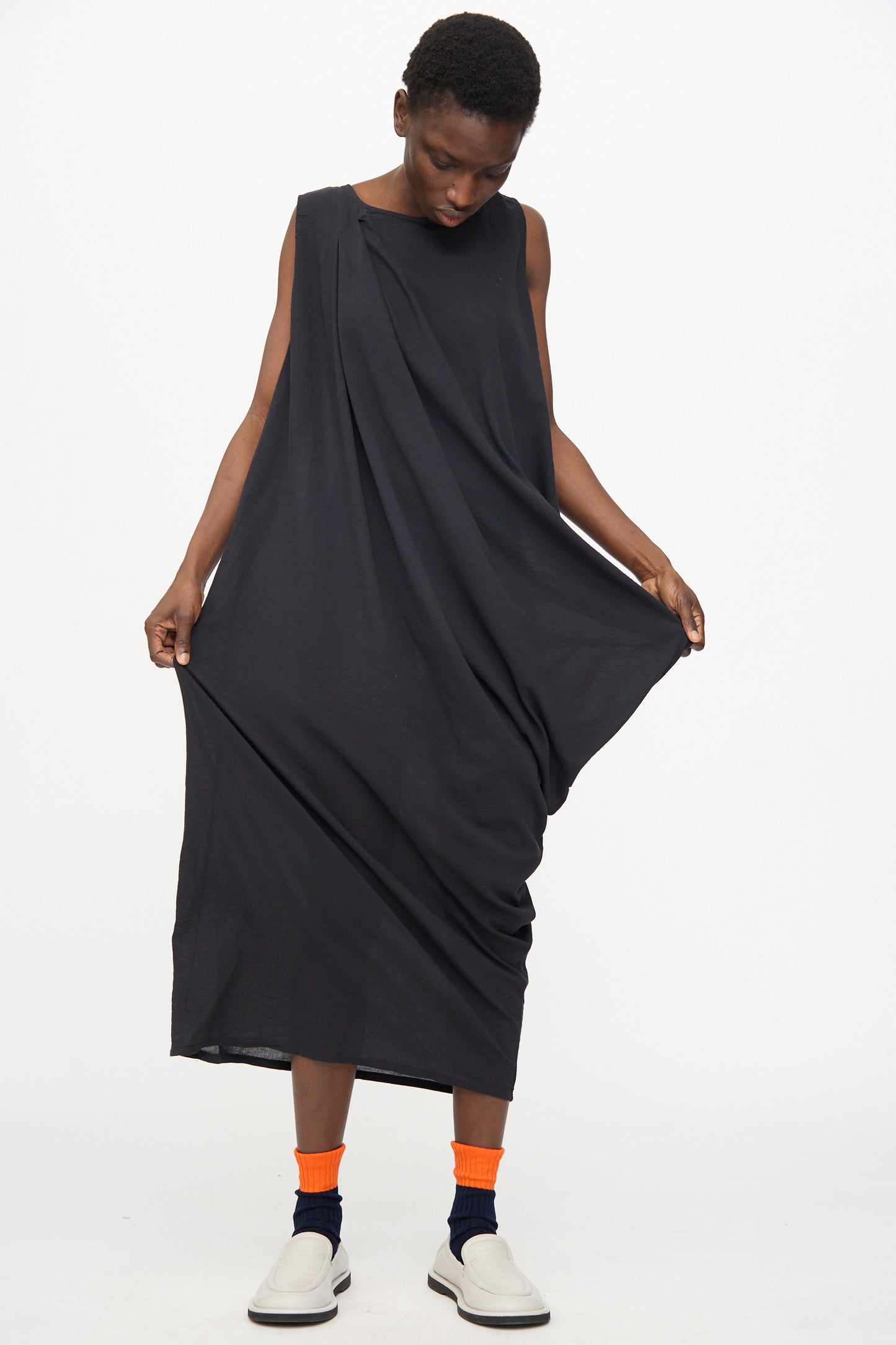 A person stands against a plain white background, looking down and holding out one side of the Black Crane Greek Column Dress in Sumi Black, styled with orange and black socks and white slip-on shoes.
