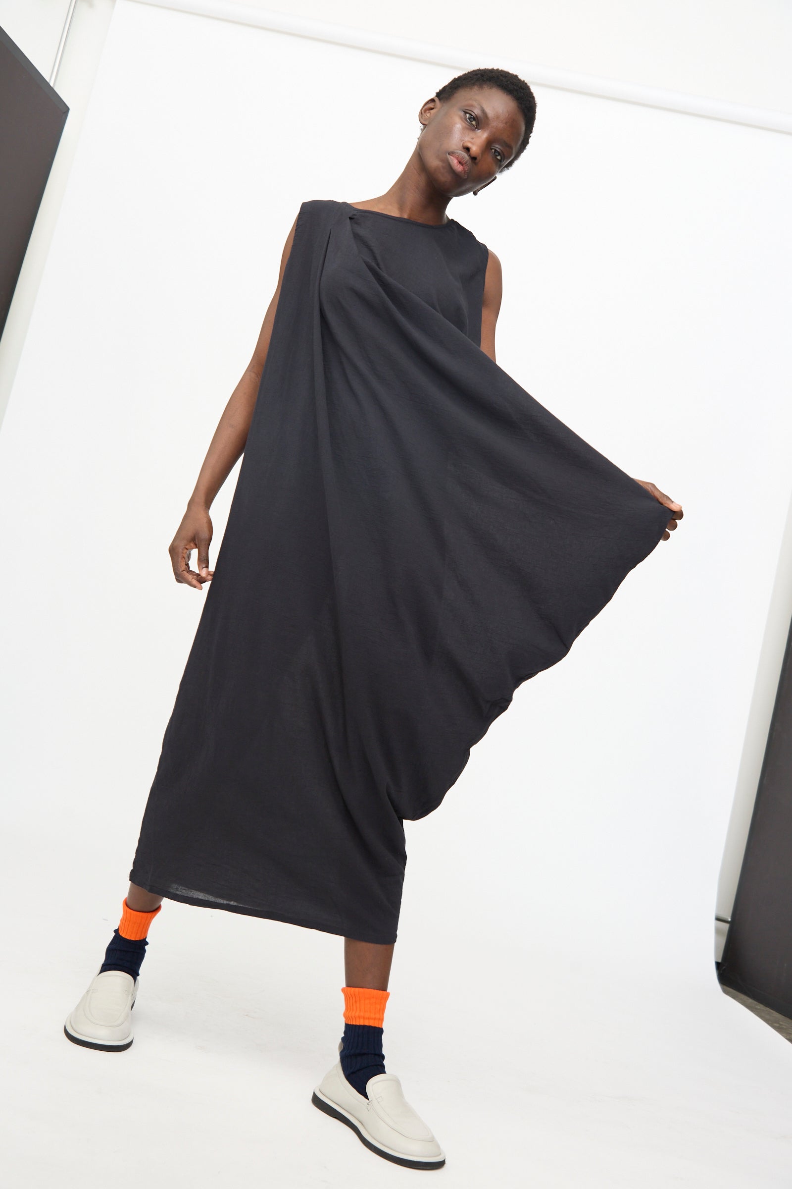A person poses in a studio wearing Black Crane’s Greek Column Dress in Sumi Black, styled with orange socks, black anklets, and white slip-on shoes, holding out one side of the loose asymmetrical dress.