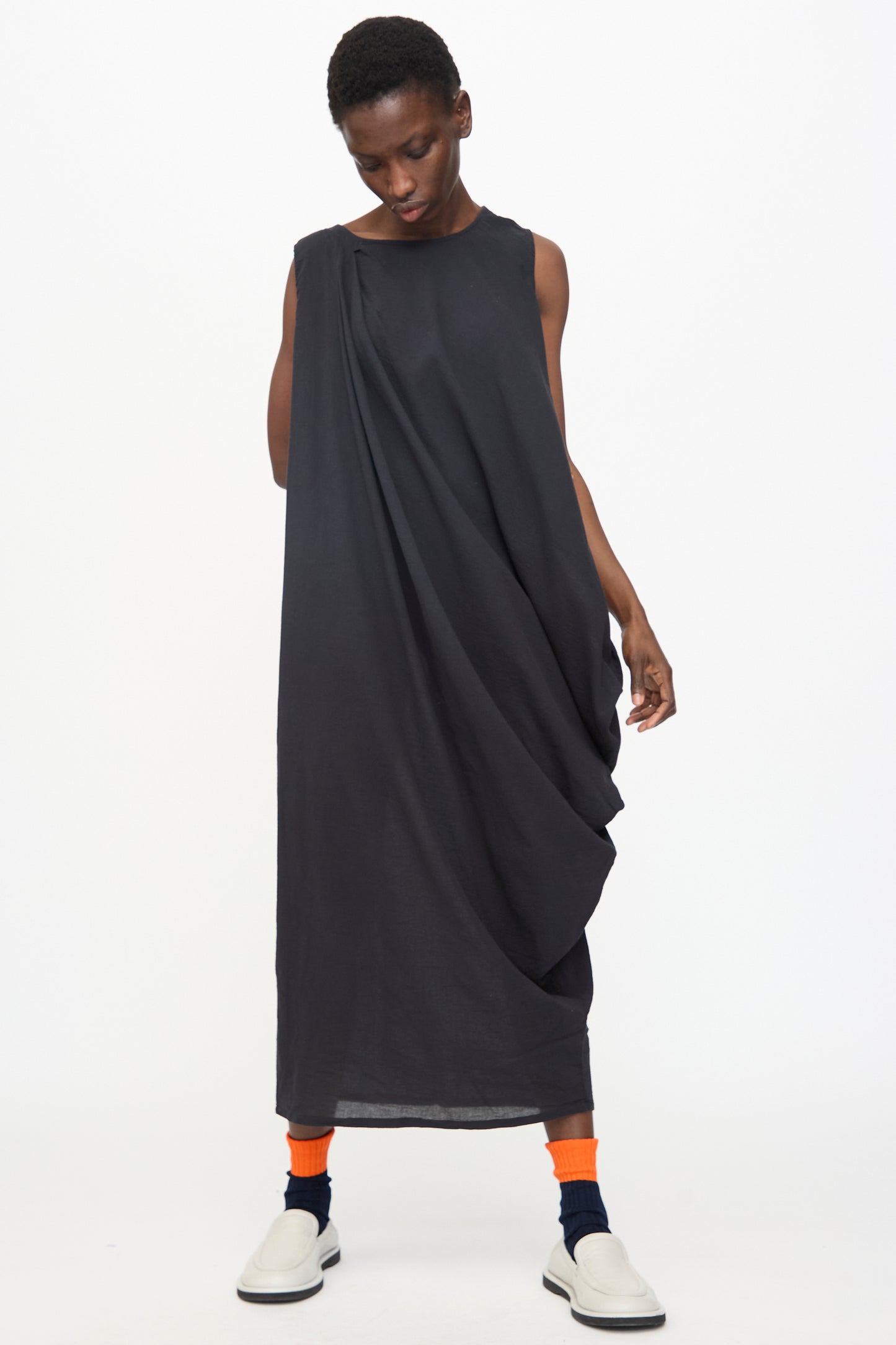 A person wears the Black Crane Greek Column Dress in Sumi Black, paired with bright orange socks, navy accents, and white slip-on shoes, standing against a plain white background.