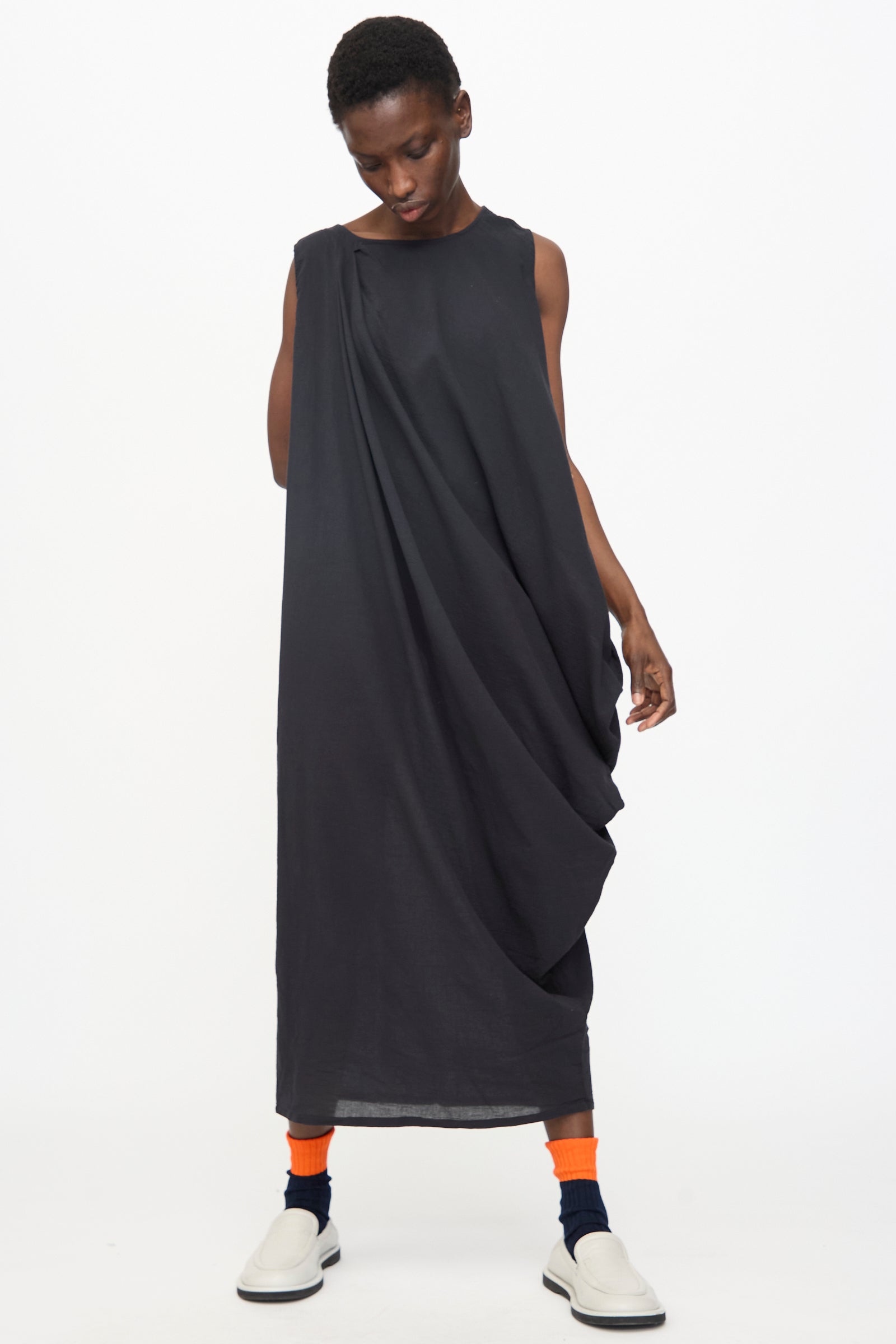 A person wears the Black Crane Greek Column Dress in Sumi Black, paired with bright orange socks, navy accents, and white slip-on shoes, standing against a plain white background.