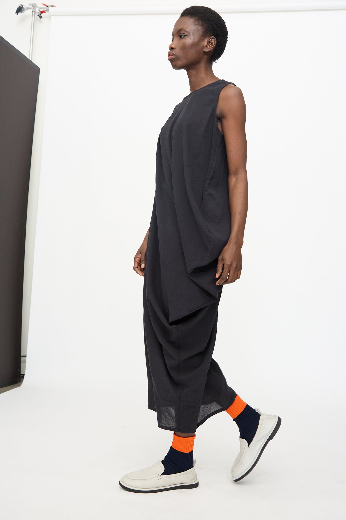 A person in the Black Crane Greek Column Dress in Sumi Black, paired with orange and navy socks and white slip-on shoes, stands against a plain white background.